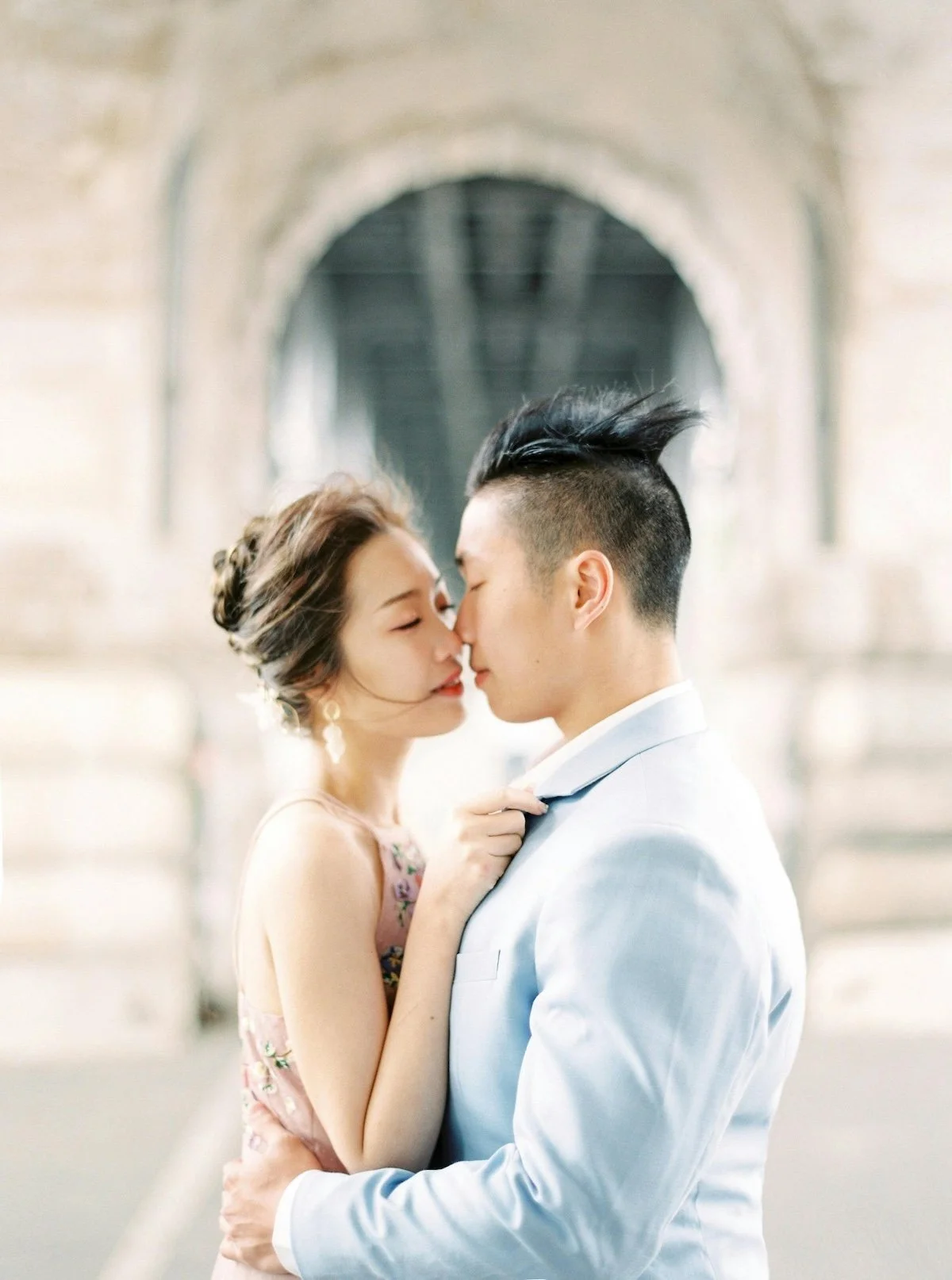 A couple dressed in formal attire sharing a kiss under an archway with a blurred background.