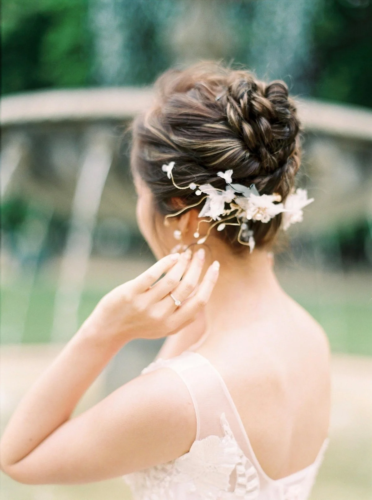 A woman with braided hair decorated with white flowers, wearing a wedding dress, is touching her neck and showing a wedding ring on her finger, with a blurred outdoor background.
