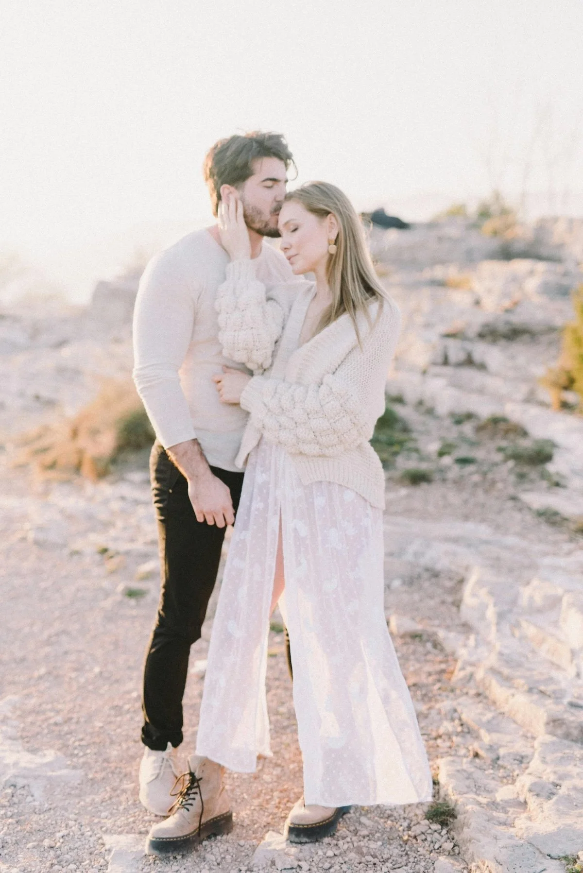 A couple standing close together outdoors during sunset, embracing each other in a desert landscape with rocks and sparse vegetation, both dressed in light clothing and wearing boots.