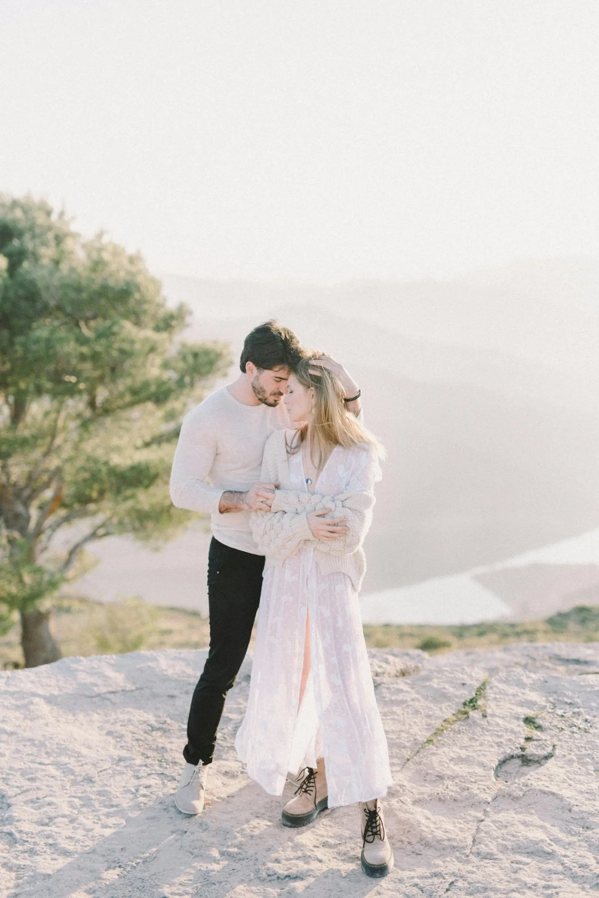 A couple standing close together outdoors on a rocky area, with a tree and sky in the background, sharing an intimate moment.