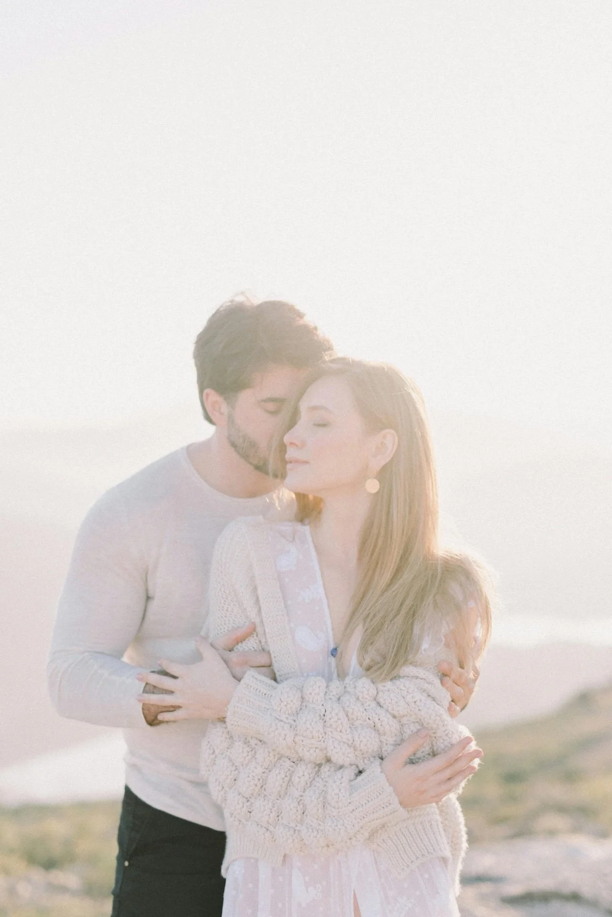 A man and woman embracing outdoors in soft sunlight. The man has dark hair and a beard, and the woman has long blonde hair, both with their eyes closed. The woman is wearing a light-colored cardigan and dress, and the man is in a light-colored shirt.