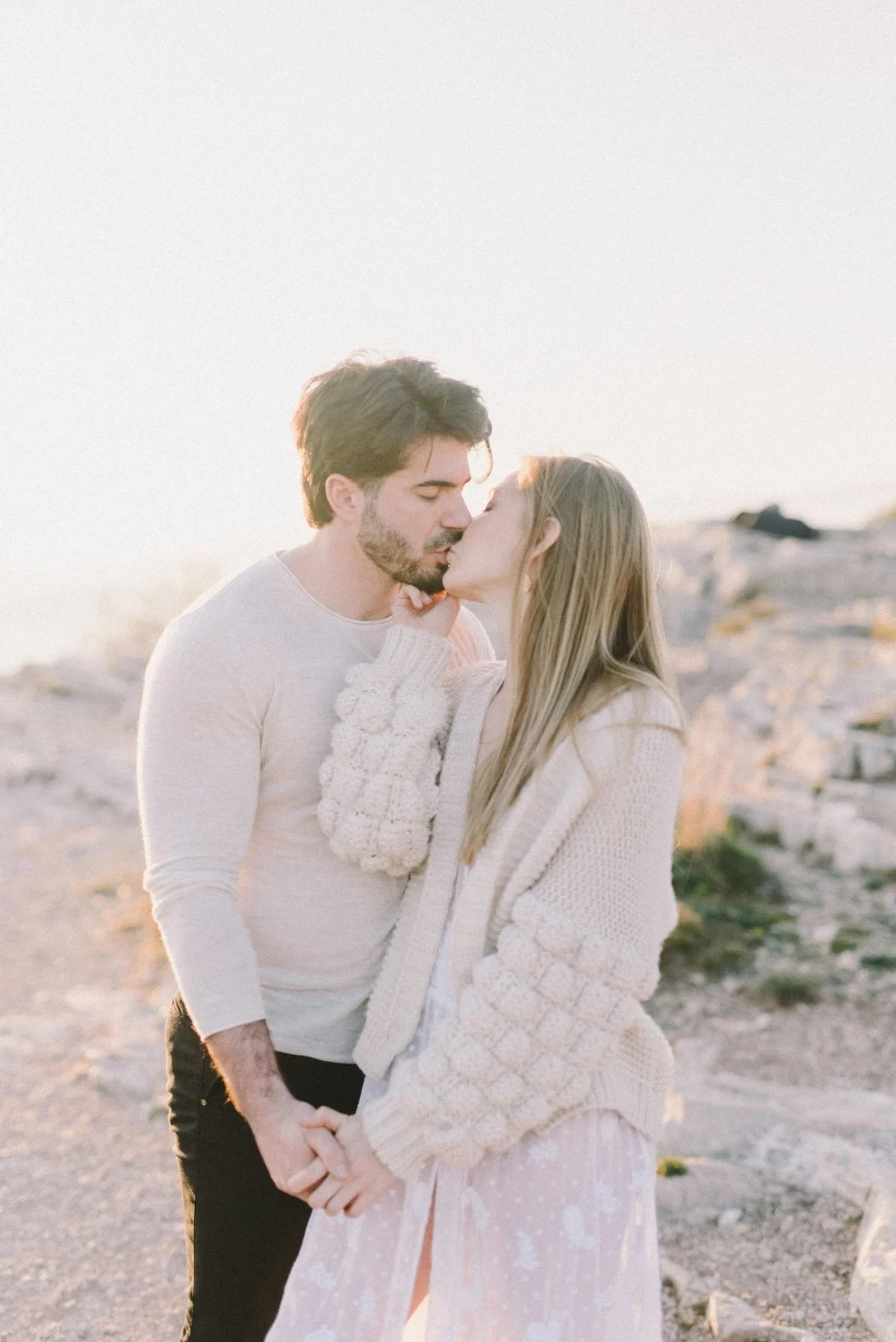 A young couple sharing a kiss outdoors during sunset, holding hands, with rocky terrain in the background.