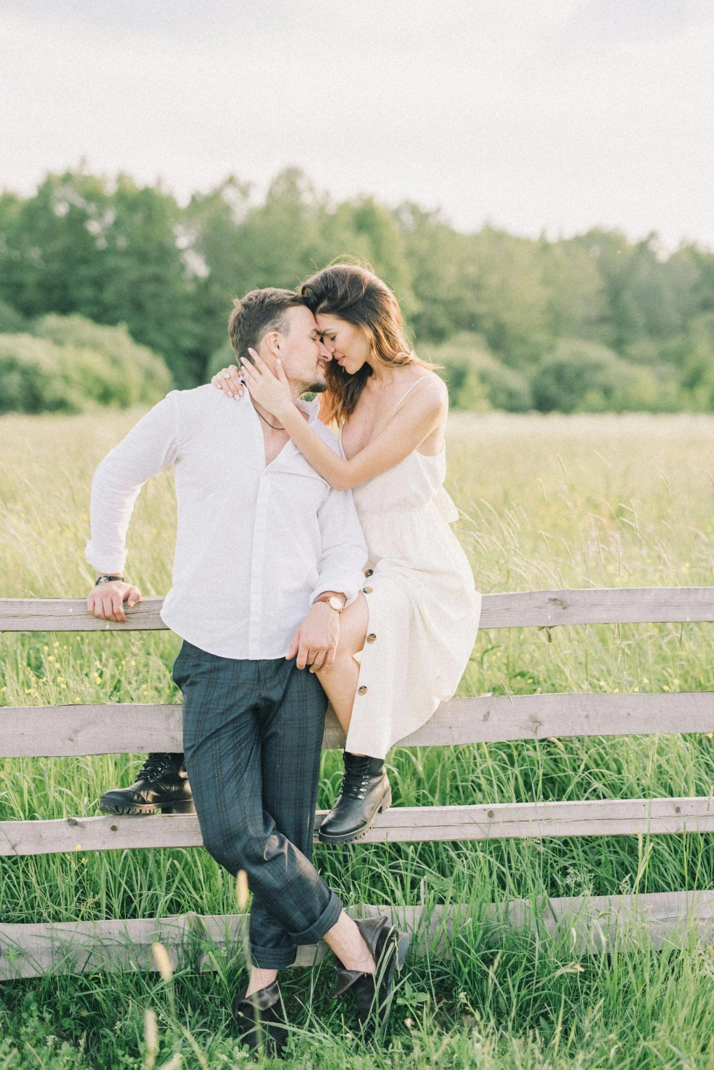 A romantic couple sits on a wooden fence in a green field, touching foreheads and smiling, with a background of trees and an open sky.
