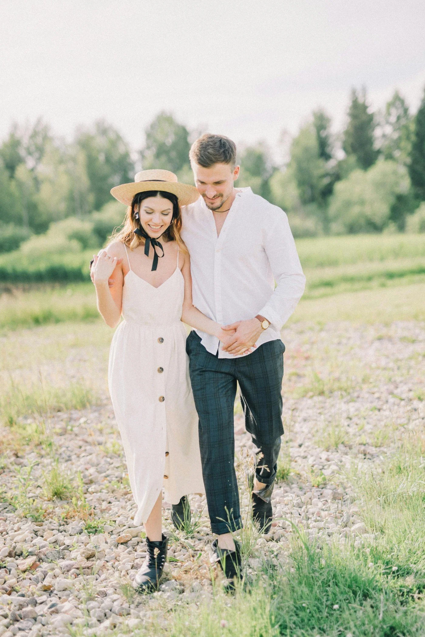 A young couple walking outdoors on a rocky trail, smiling and looking at each other, with trees in the background during daytime.