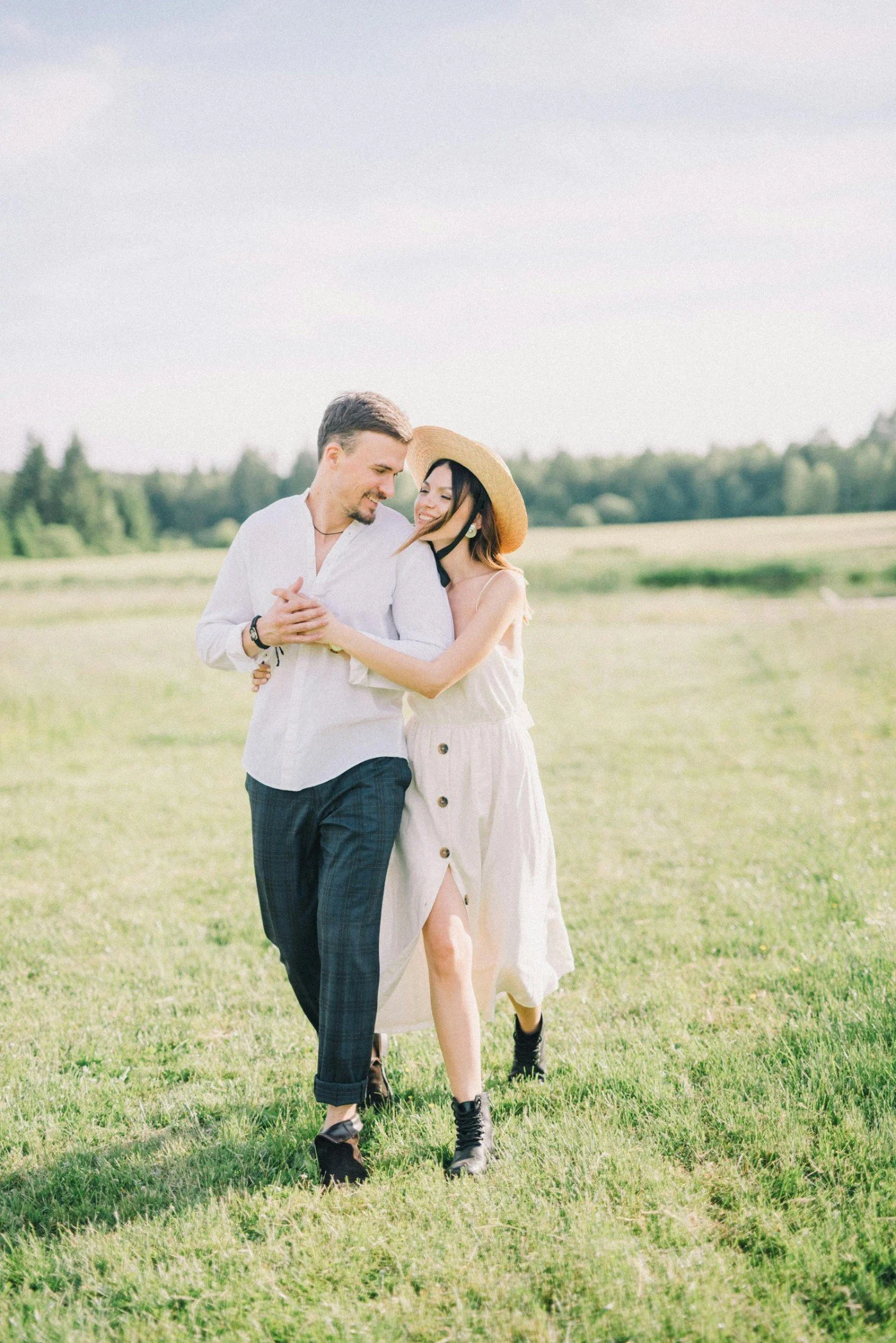 A man and woman smiling and walking together in a grassy field, with trees in the background. The woman wears a straw hat and a light-colored dress, and the man has short dark hair, a beard, and wears a white shirt with rolled-up sleeves and dark pan