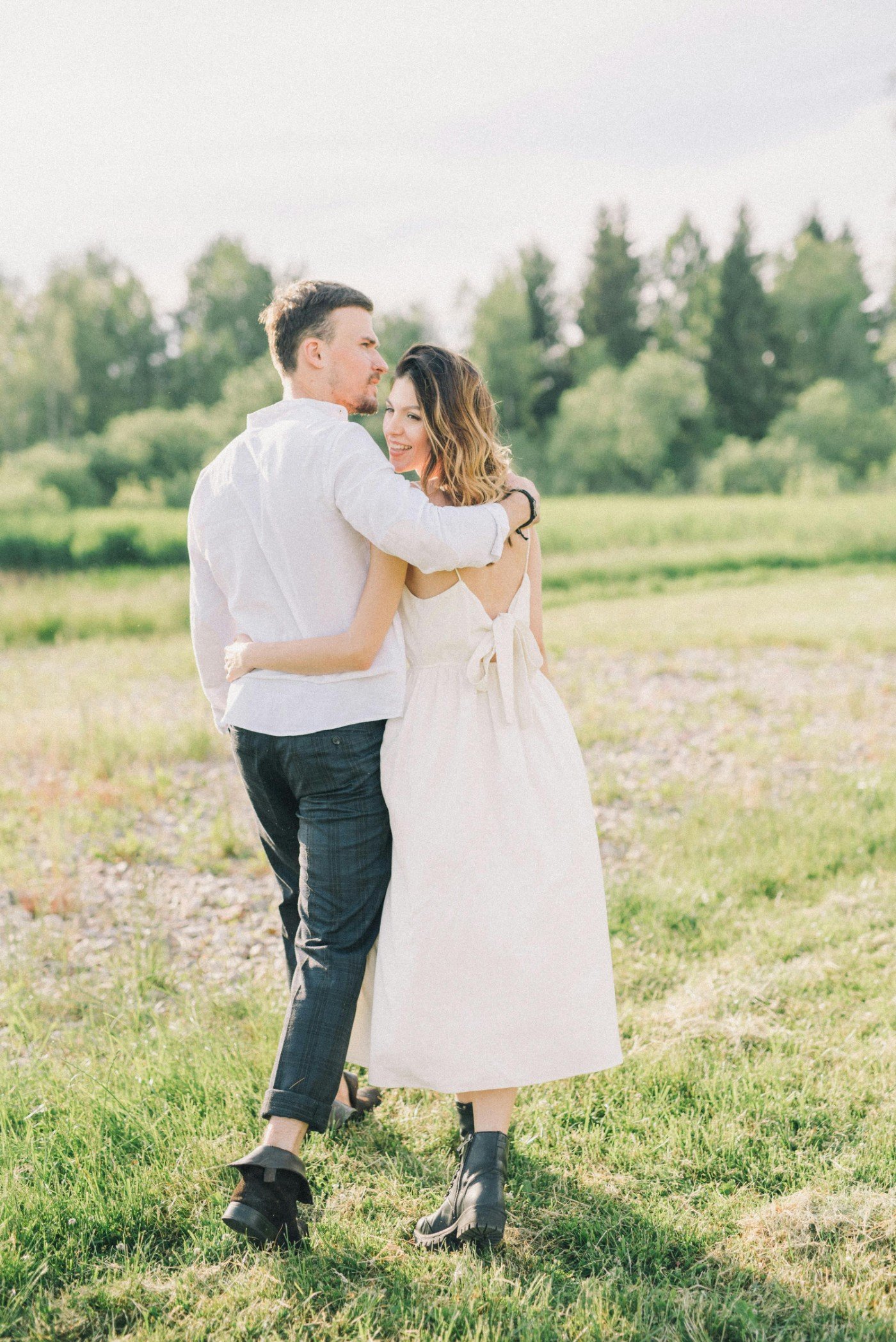 A couple hugging outdoors in a grassy field with trees in the background, both smiling and looking happy, wearing casual clothing.