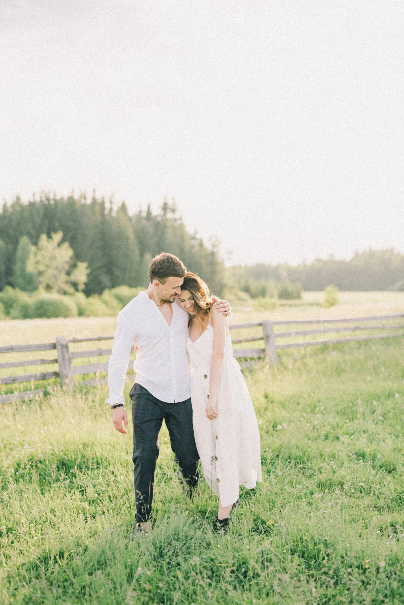 A young couple walking in a grassy field with a wooden fence, surrounded by green trees and hills, during daytime, sharing a tender moment.
