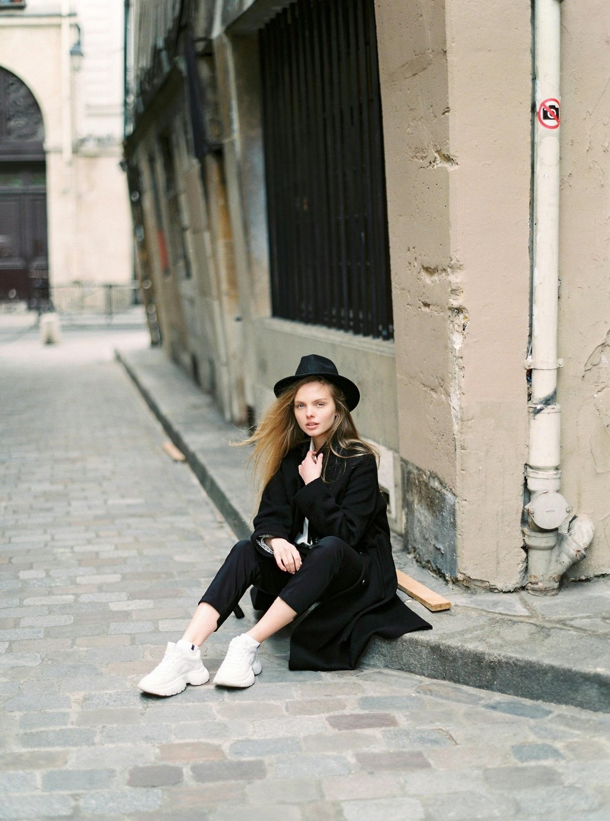 A young woman with long blonde hair, wearing a black hat and black coat, sits on a sidewalk against a beige building wall. She is wearing white sneakers, and her hair is blowing to the side. The building has a barred window, a white pipe with a no-sm