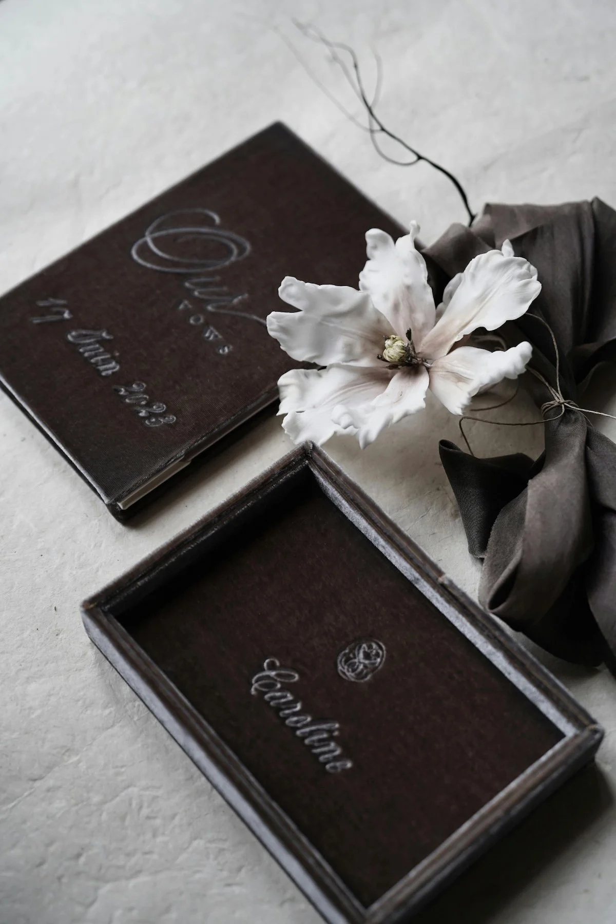 A white flower with large petals and a small bud, placed on a gray wrapped cloth, beside a small jewelry box and a jewelry booklet, all on a light surface.