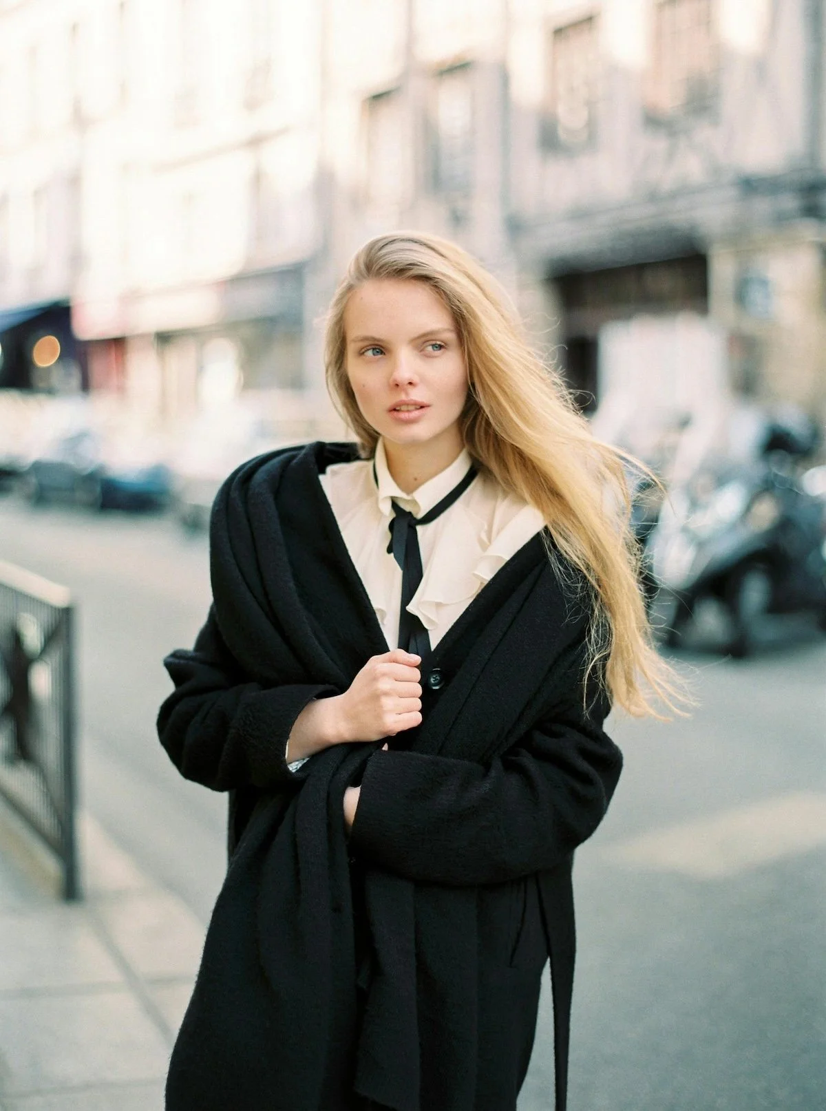 A young woman with long blonde hair and blue eyes standing on a city sidewalk, wearing a white blouse with ruffles, a black coat draped over her shoulders, and a black tie, with a blurred urban street background.