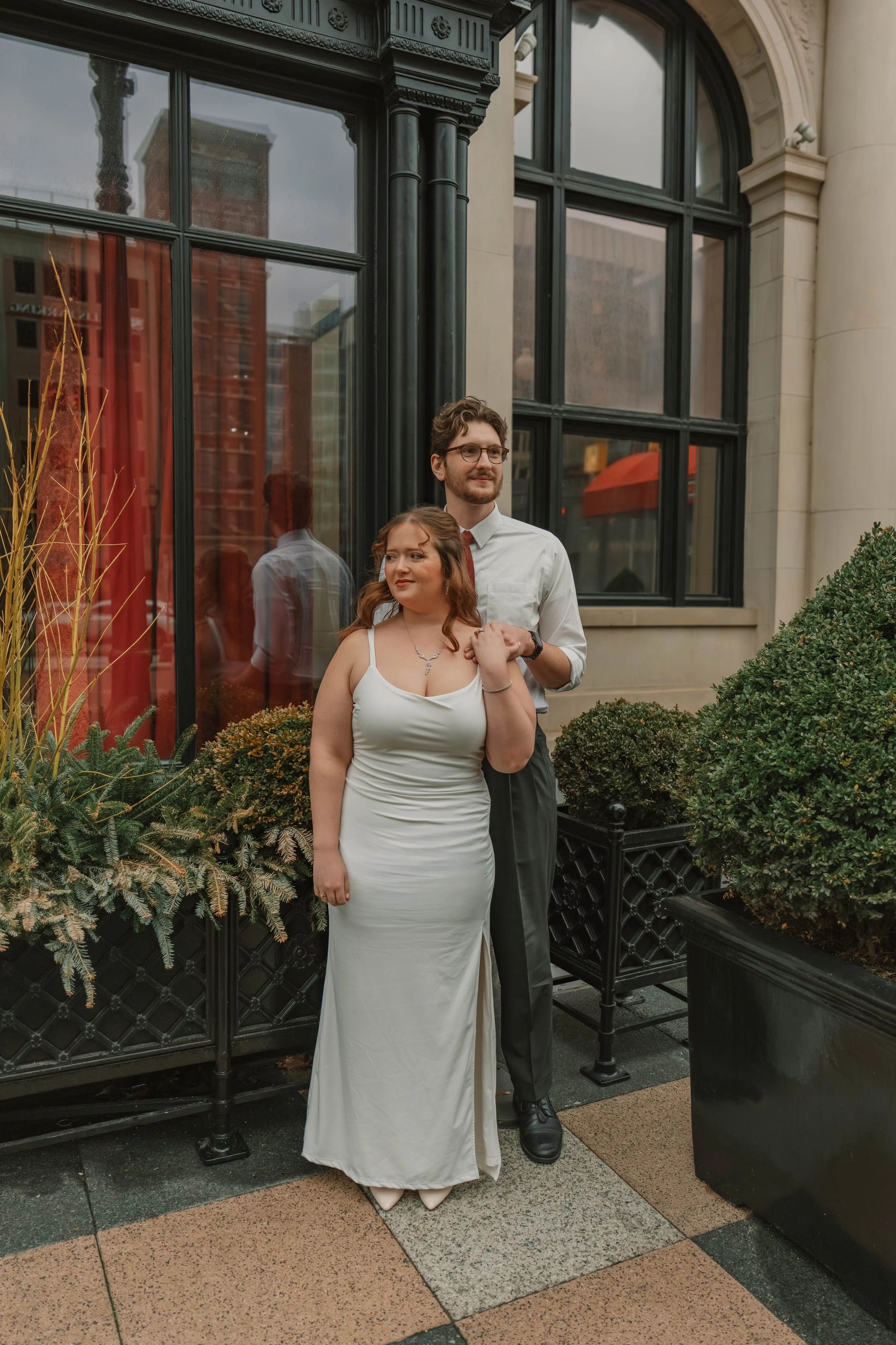 A couple standing outside a building with large windows, surrounded by potted plants, in formal wedding attire. The woman wears a white dress and the man wears a white shirt with a red tie and gray trousers.
