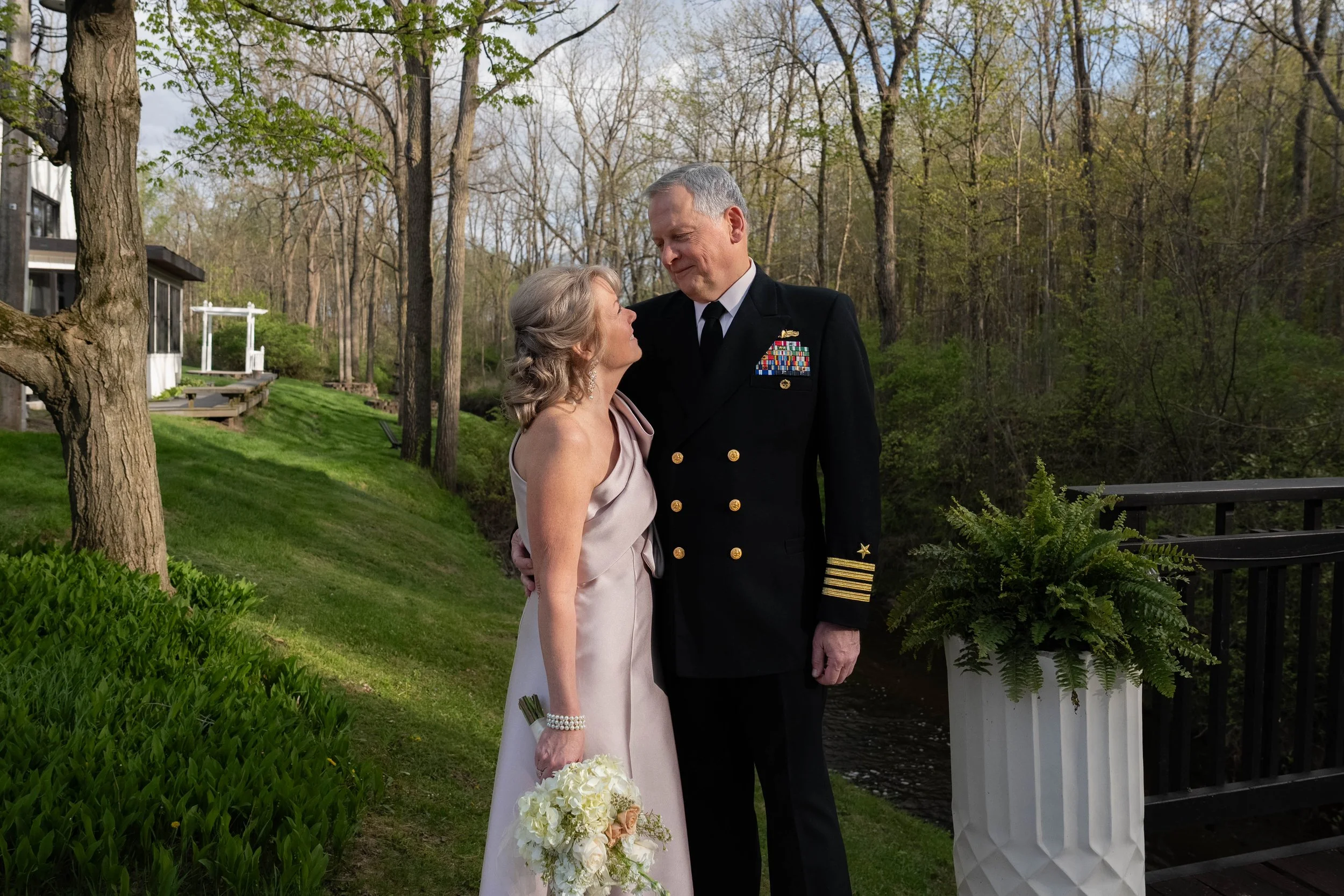 A couple in wedding attire standing outdoors near water, with trees and a grassy area surrounding them, during daytime.