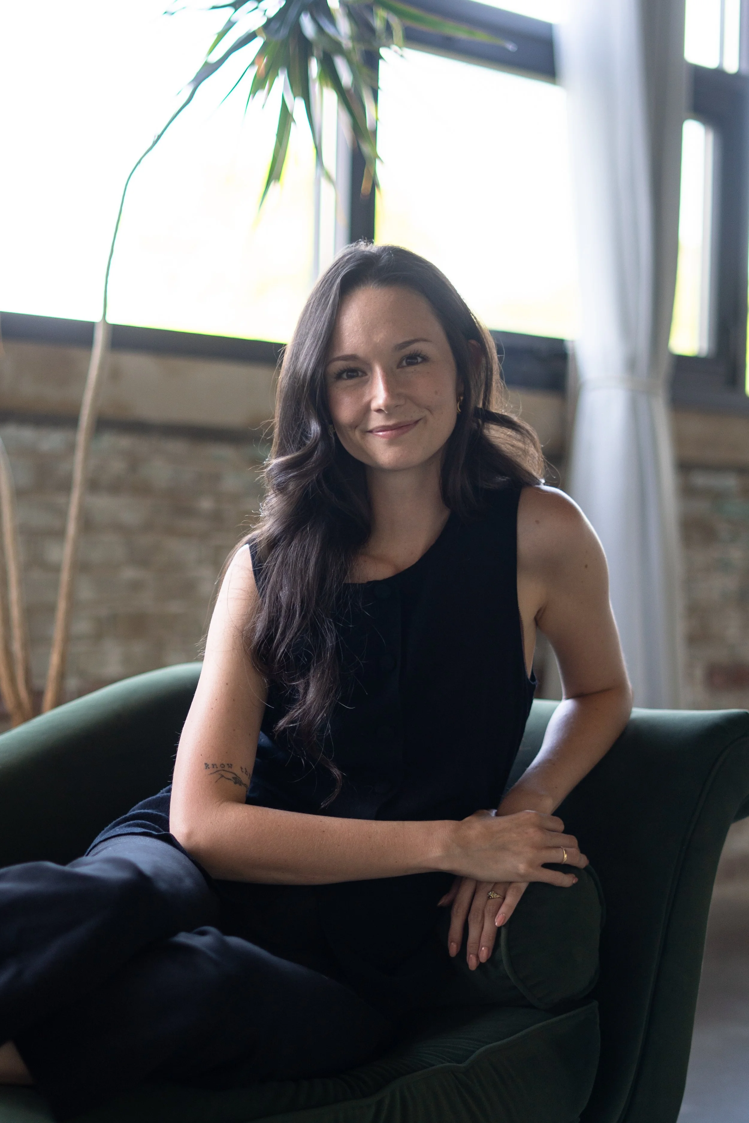 Young woman with long dark hair sitting on a dark green velvet sofa, smiling slightly, with large windows and a tall indoor plant in the background, in bright natural light.