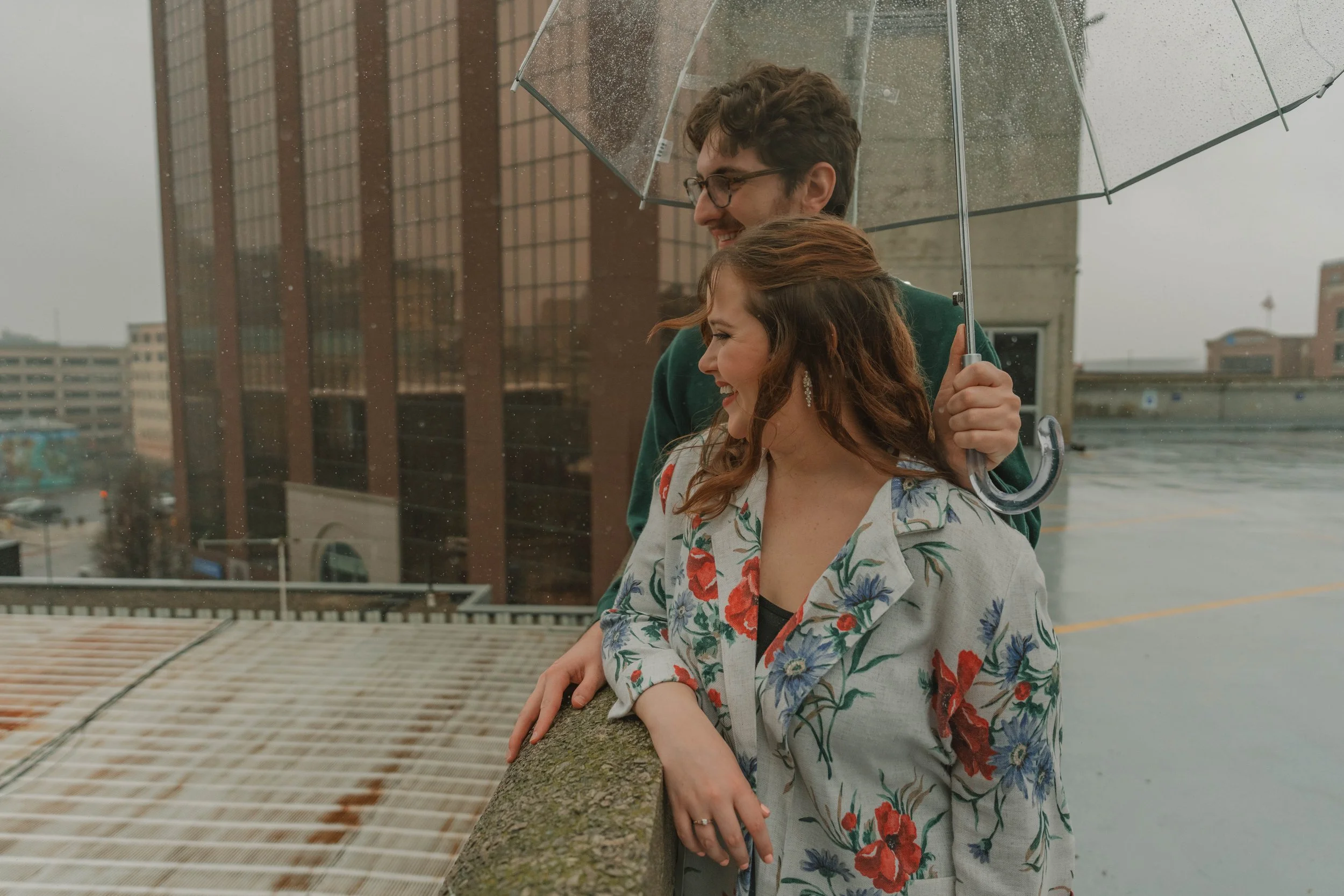 A man and woman standing under a transparent umbrella on a rainy day. The man is smiling and wearing glasses, the woman is leaning her head on his shoulder, both are smiling, with a building and cityscape in the background.