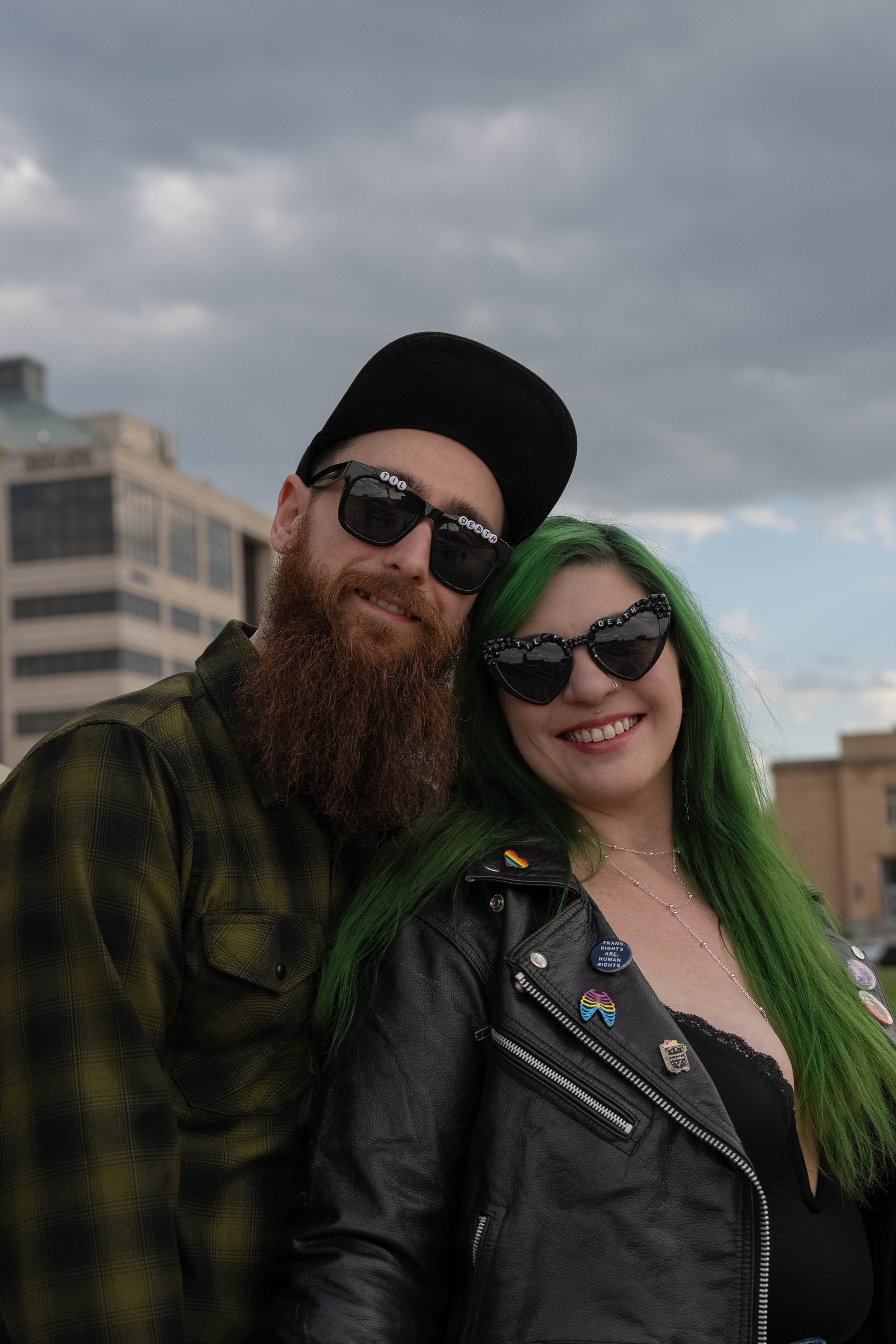 Two people smiling outdoors on a cloudy day, with a city building in the background. The person on the left has a beard, sunglasses, a black cap, and a green plaid shirt. The person on the right has bright green hair, heart-shaped sunglasses with the