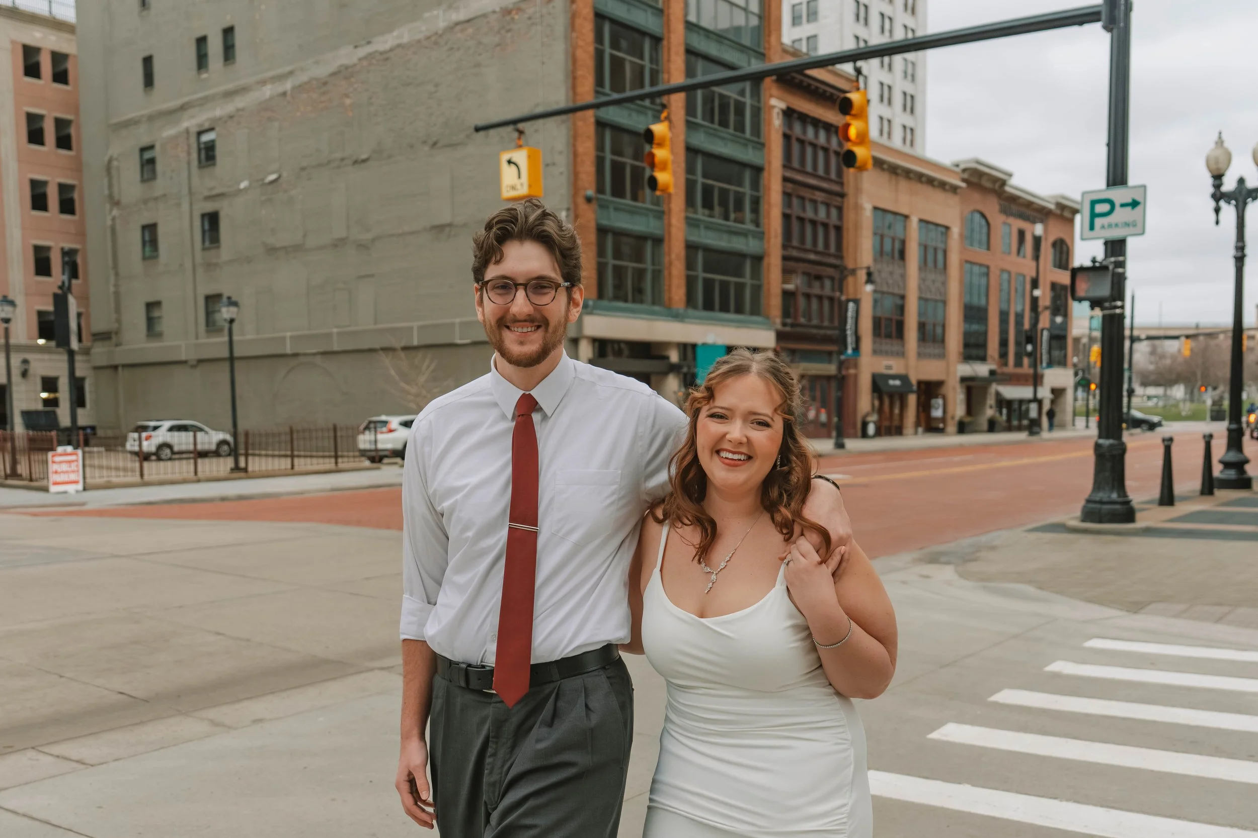 Smiling man in dress shirt, tie, and glasses standing next to woman in white dress, on city street with buildings and traffic lights in the background.