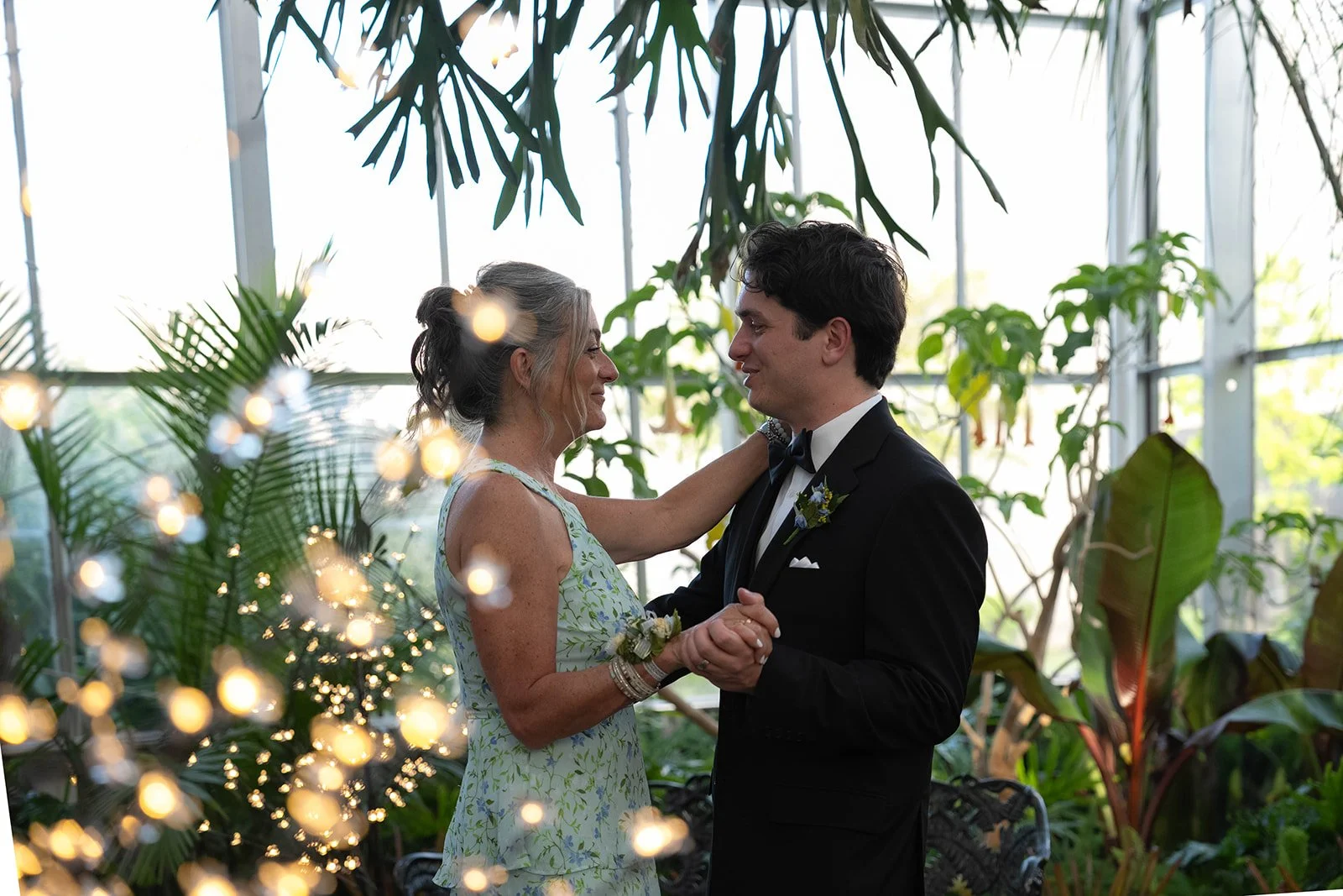 A woman and a man are dancing closely in a lush greenhouse or conservatory, surrounded by green plants and warm lighting, during a wedding or special occasion.