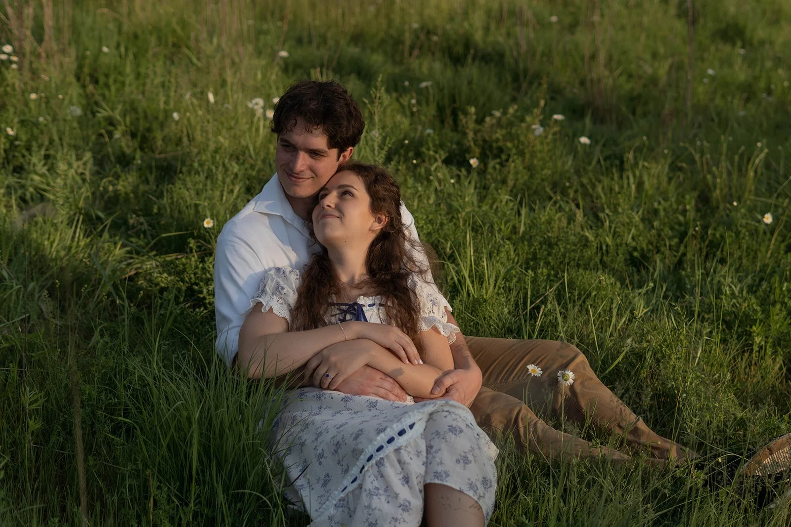 A young couple sitting in a grassy field with small white flowers, embracing each other and gazing into each other's eyes, evoking a romantic mood.