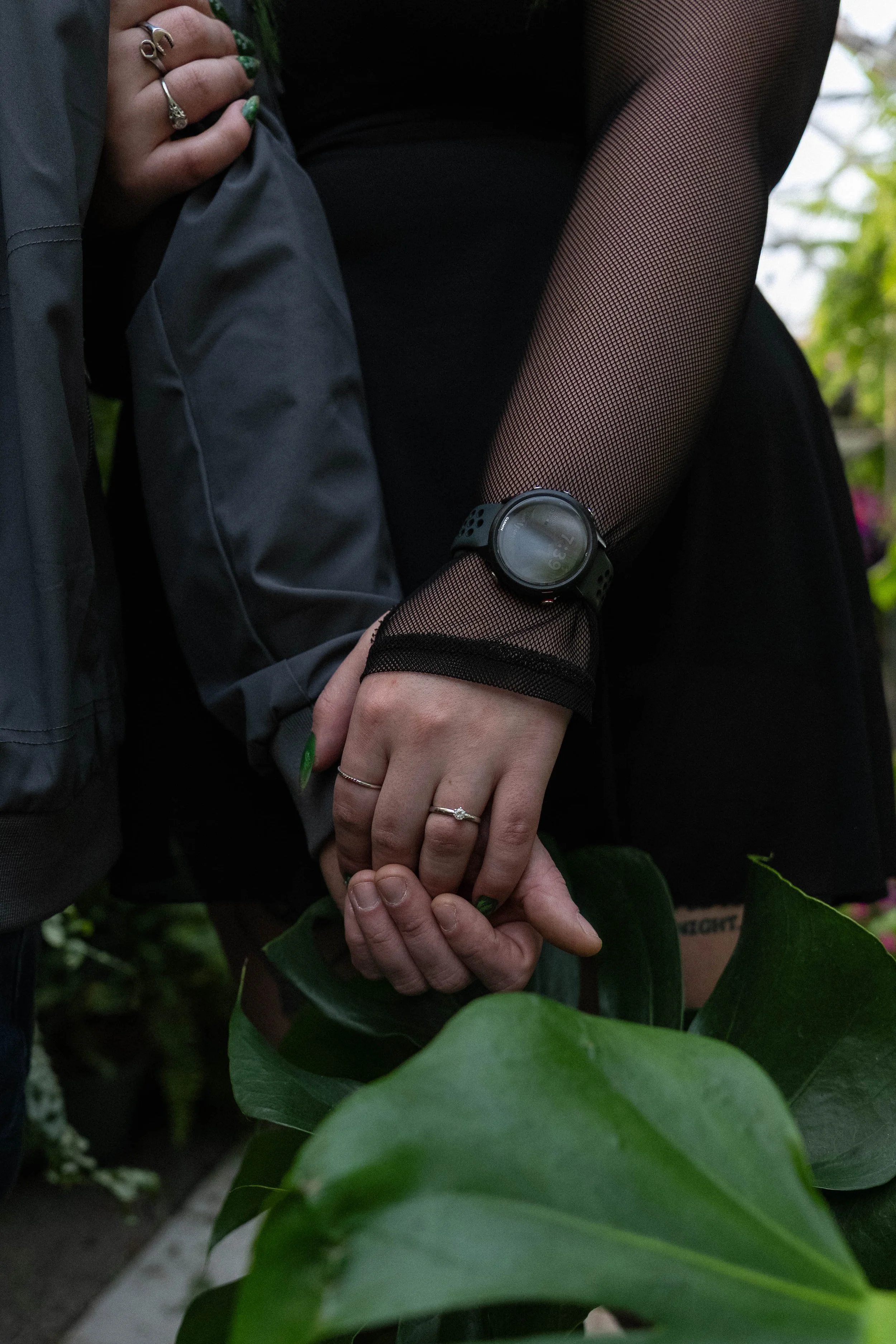 Two people holding hands among green plants, one wearing a smartwatch and fishnet stockings.