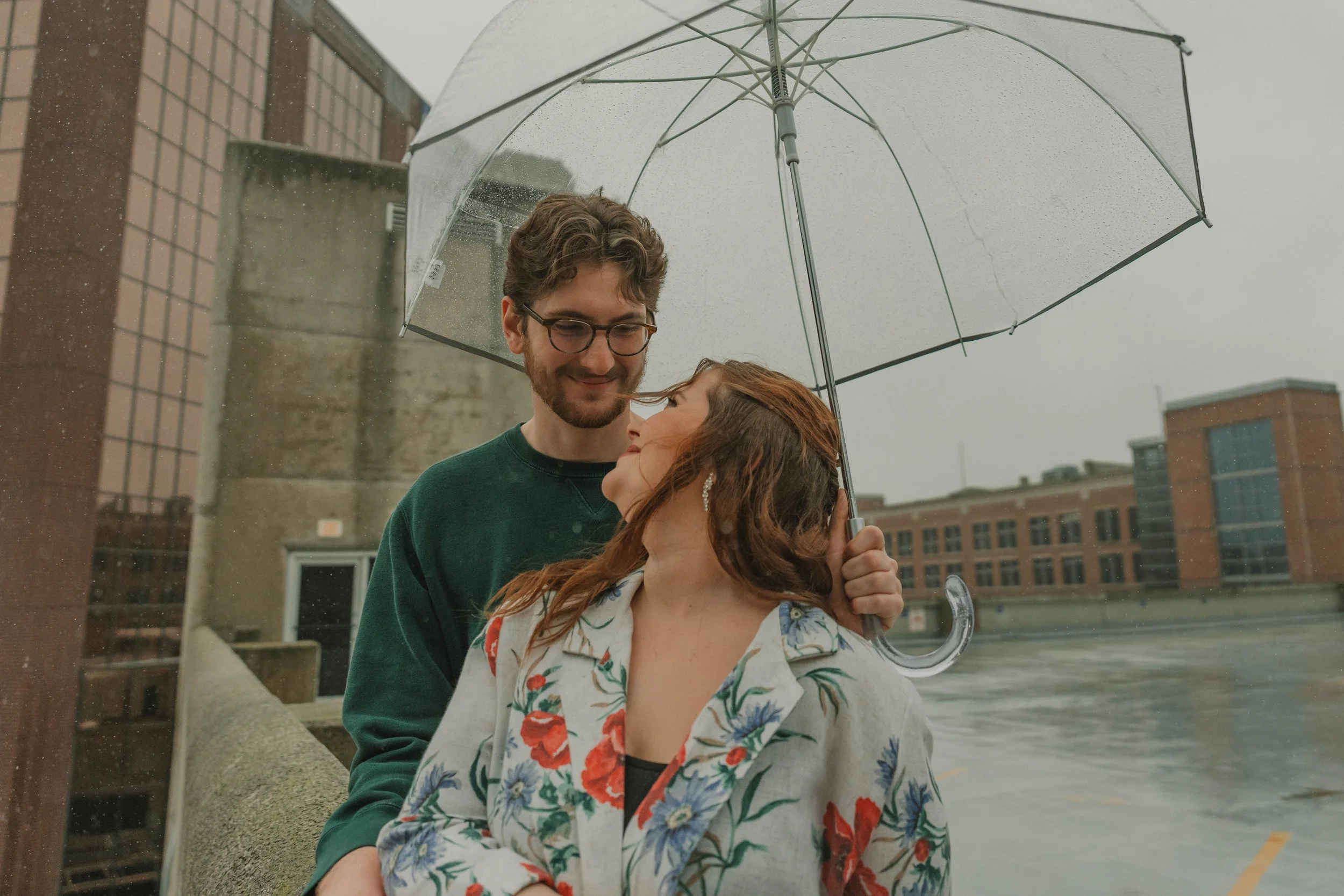 A young man and woman standing under a clear umbrella on a rainy day near a river or canal with buildings in the background.