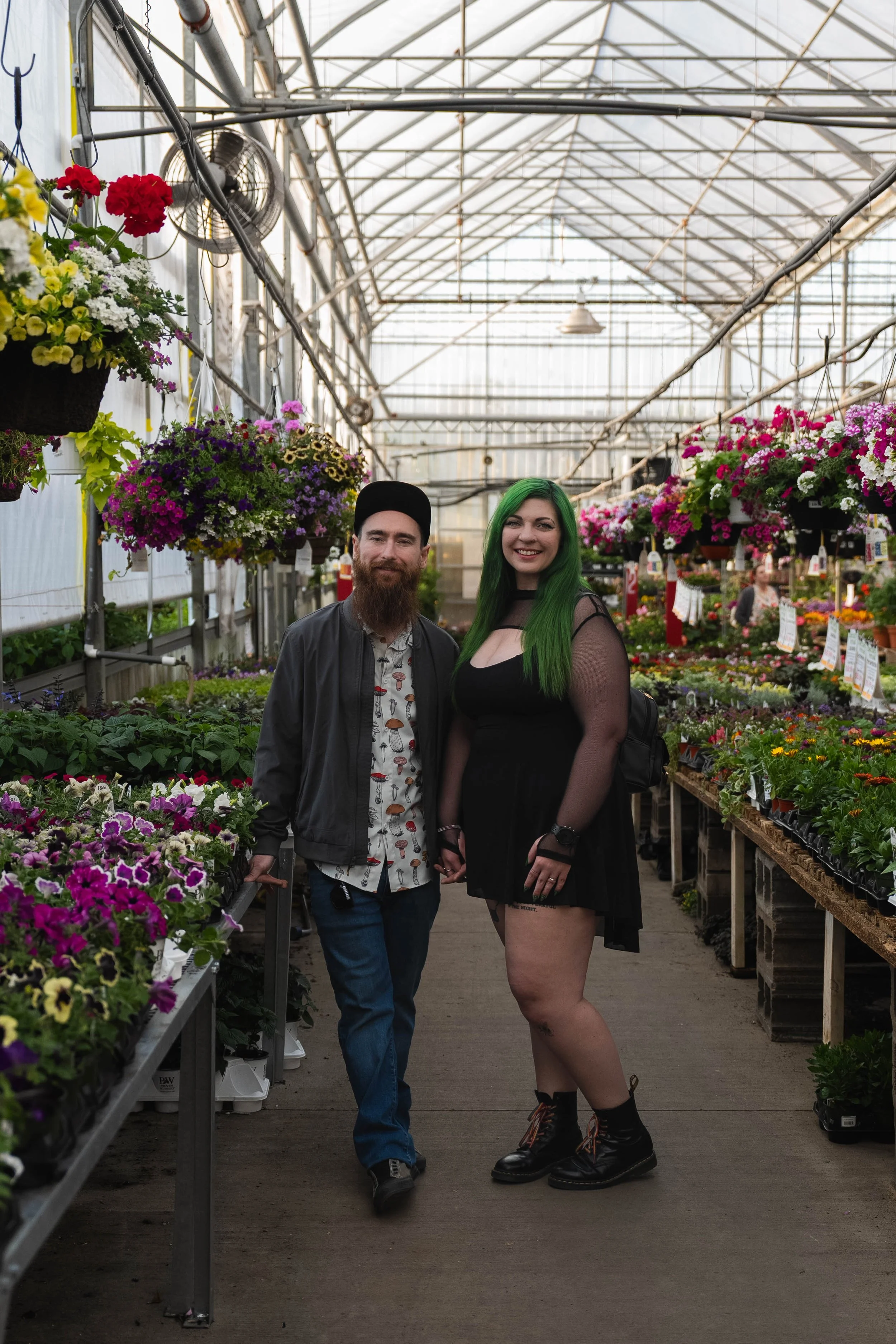 A man with a beard and a woman with green hair standing in a greenhouse surrounded by colorful flowers, both smiling at the camera.