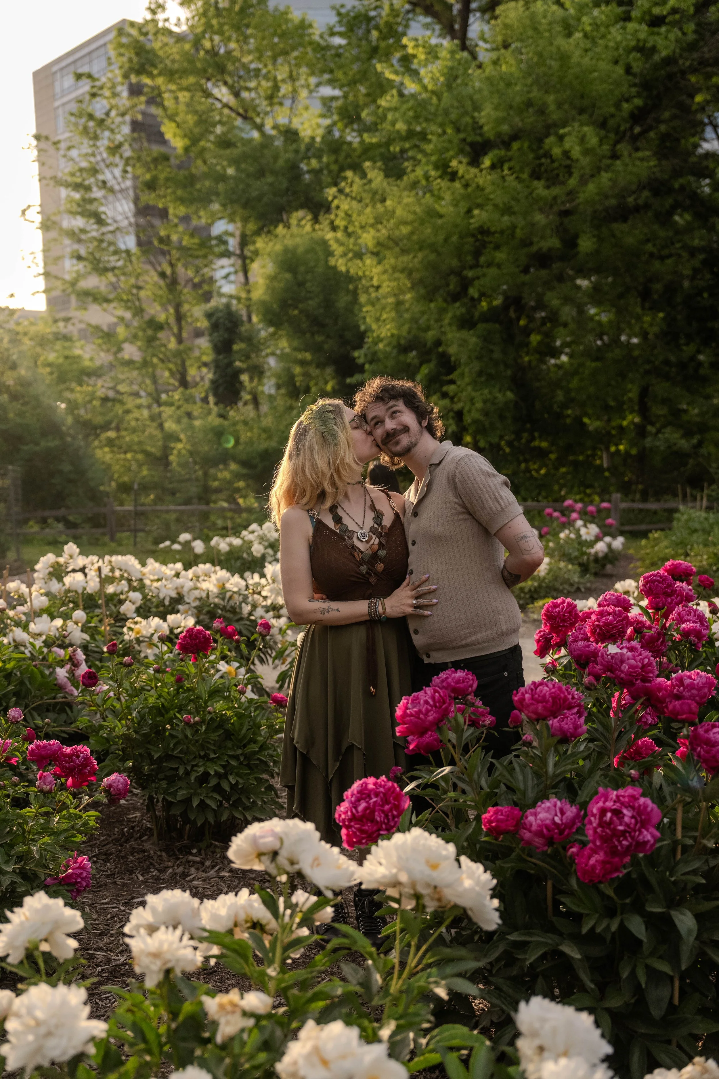 A happy couple stands in a flower garden, sharing a kiss and smiling. The woman has blonde hair and is wearing a dark green dress with jewelry; the man has curly hair and a beard, wearing a light brown shirt. The garden is filled with pink and white 