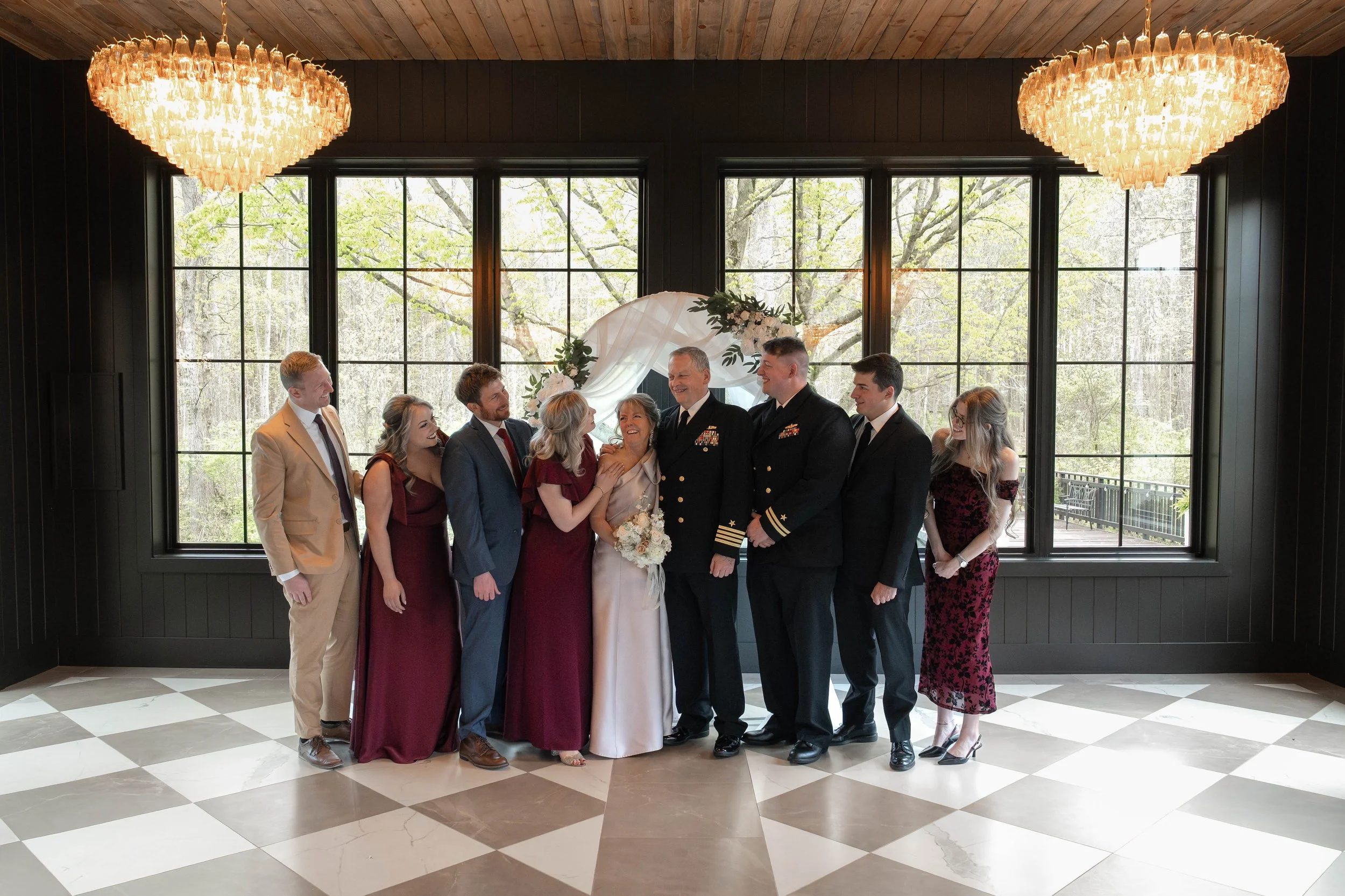 A group of nine people, including two in military uniforms and others in formal attire, standing indoors near large windows with verdant trees outside. There are two large chandeliers hanging from the ceiling and a floral wedding arch behind the grou