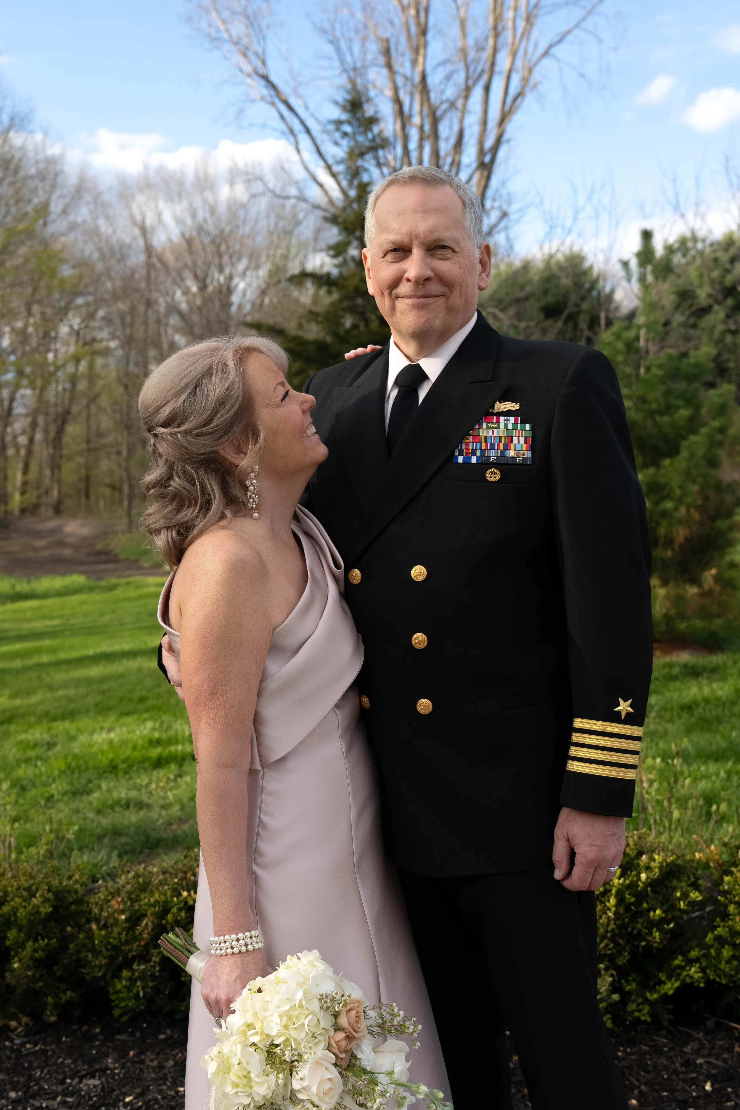 A smiling middle-aged woman in a pale pink evening gown holding a bouquet of white and blush roses, wearing pearl earrings and bracelet, standing outdoors next to a tall man in a black military dress uniform with medals, ribbons, and gold insignia, a