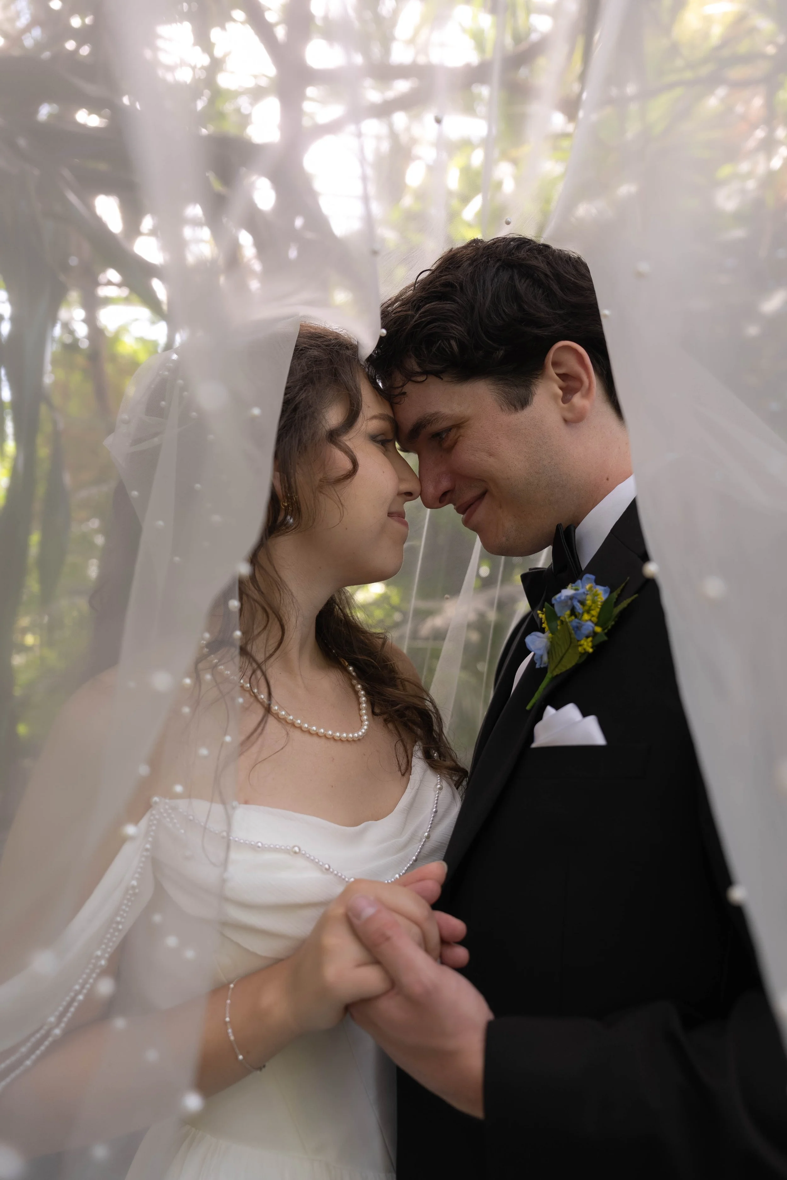 A newlywed couple, a woman in a white wedding dress and pearl necklace and a man in a tuxedo with a floral boutonniere, standing closely with foreheads touching, holding hands under a sheer veil outdoors surrounded by trees.