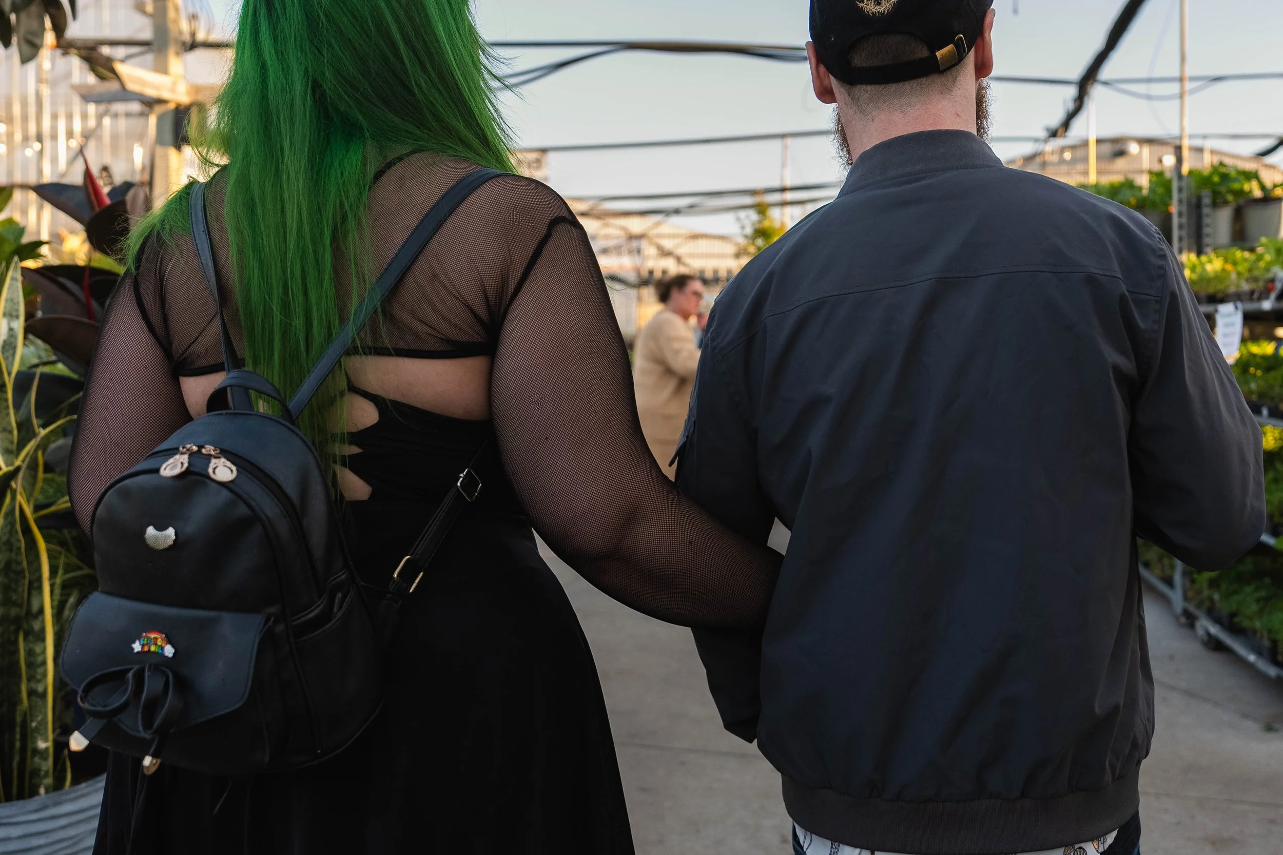 Two people with green hair and a black backpack, and a man with a beard wearing a cap, standing near plants in what appears to be a greenhouse or garden center.