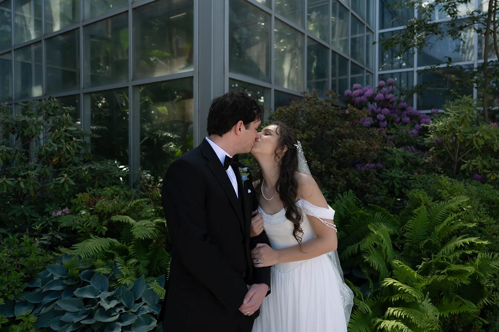 A bride and groom sharing a kiss outdoors, surrounded by lush plants and a greenhouse structure.