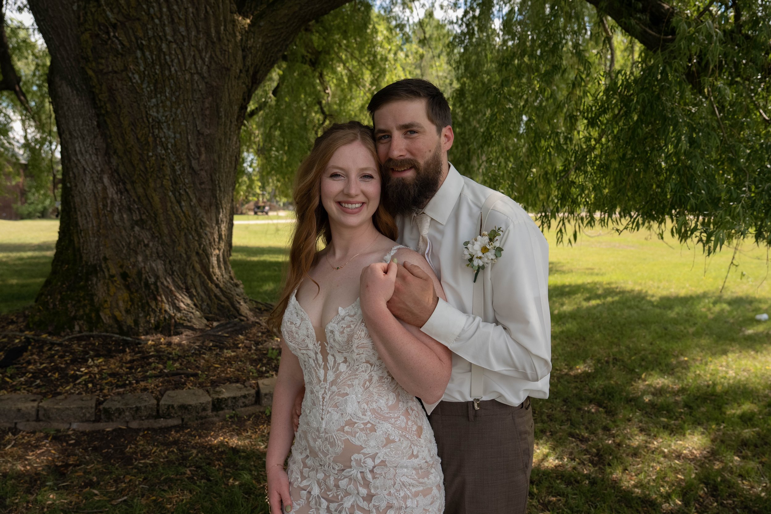 A smiling bride and groom standing outdoors beneath a large tree, with the bride wearing a lace wedding dress and the groom in a white shirt and tie, captured during daytime in a park.