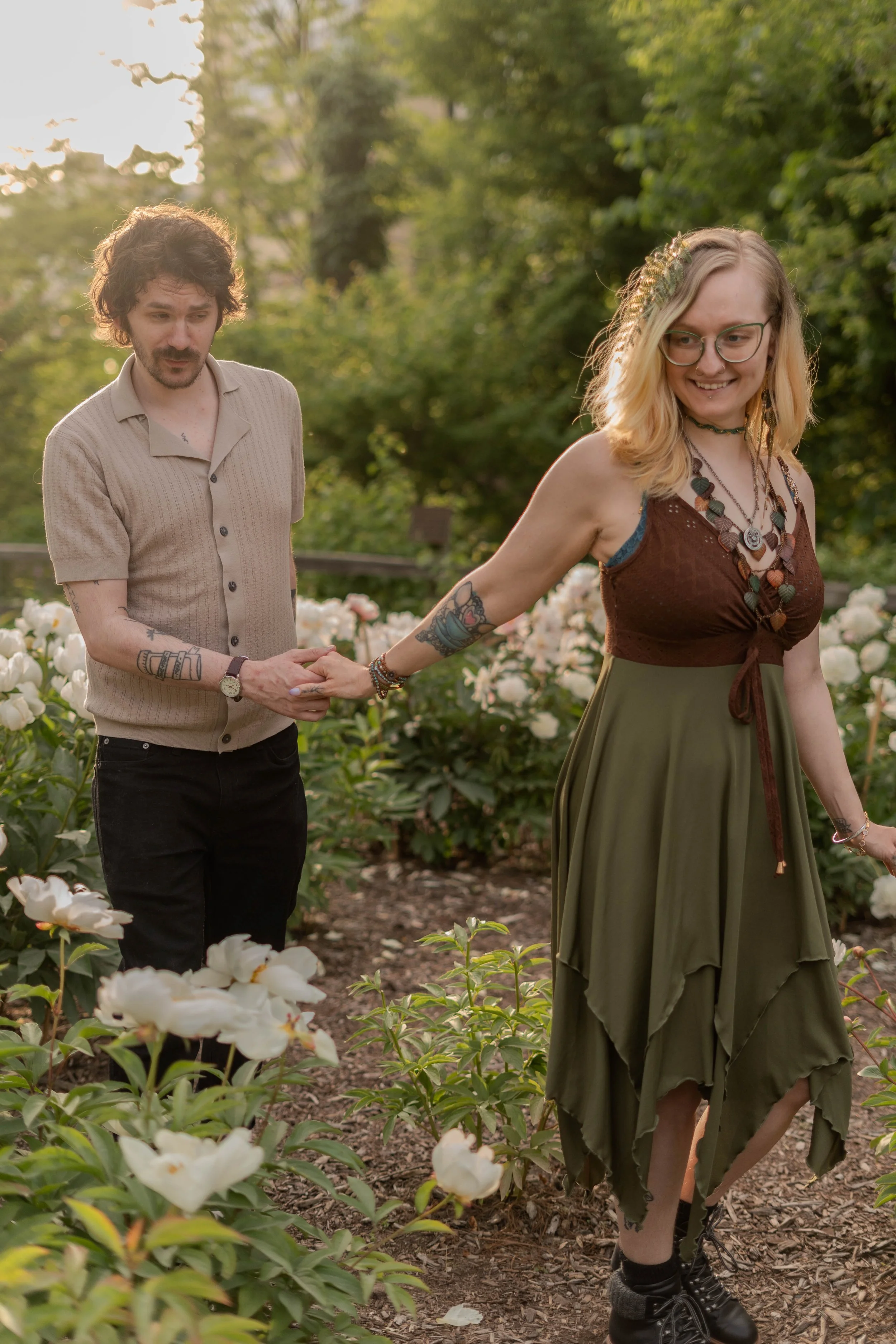 A man and a woman holding hands in a garden surrounded by white flowers and green trees, with the woman smiling and wearing glasses, multiple necklaces, and a dress, while the man has tattoos and facial hair.