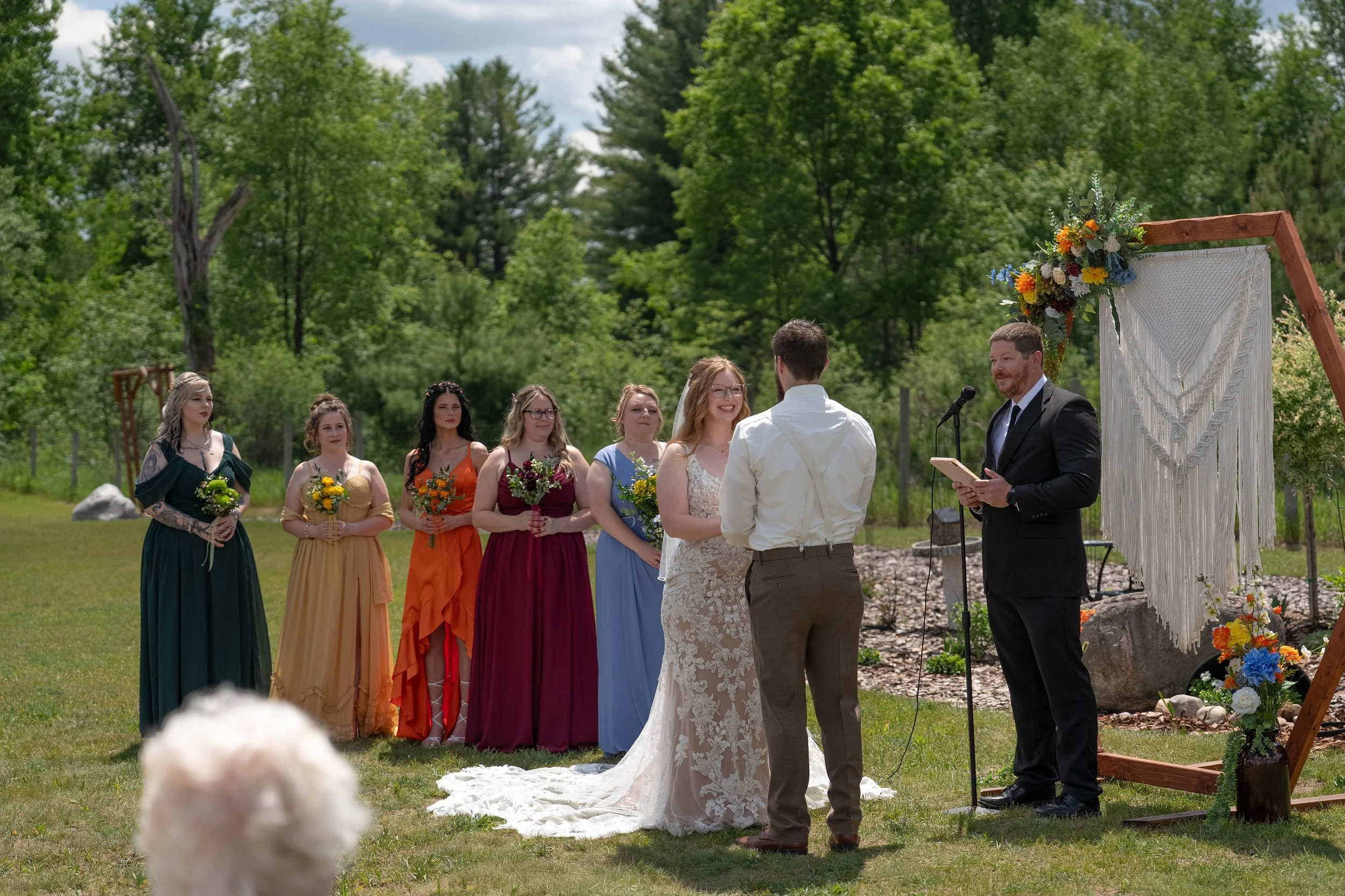 An outdoor wedding ceremony with a bride and groom standing before a officiant, surrounded by bridesmaids and groomsmen. The setting features green trees and a decorated arch with flowers.