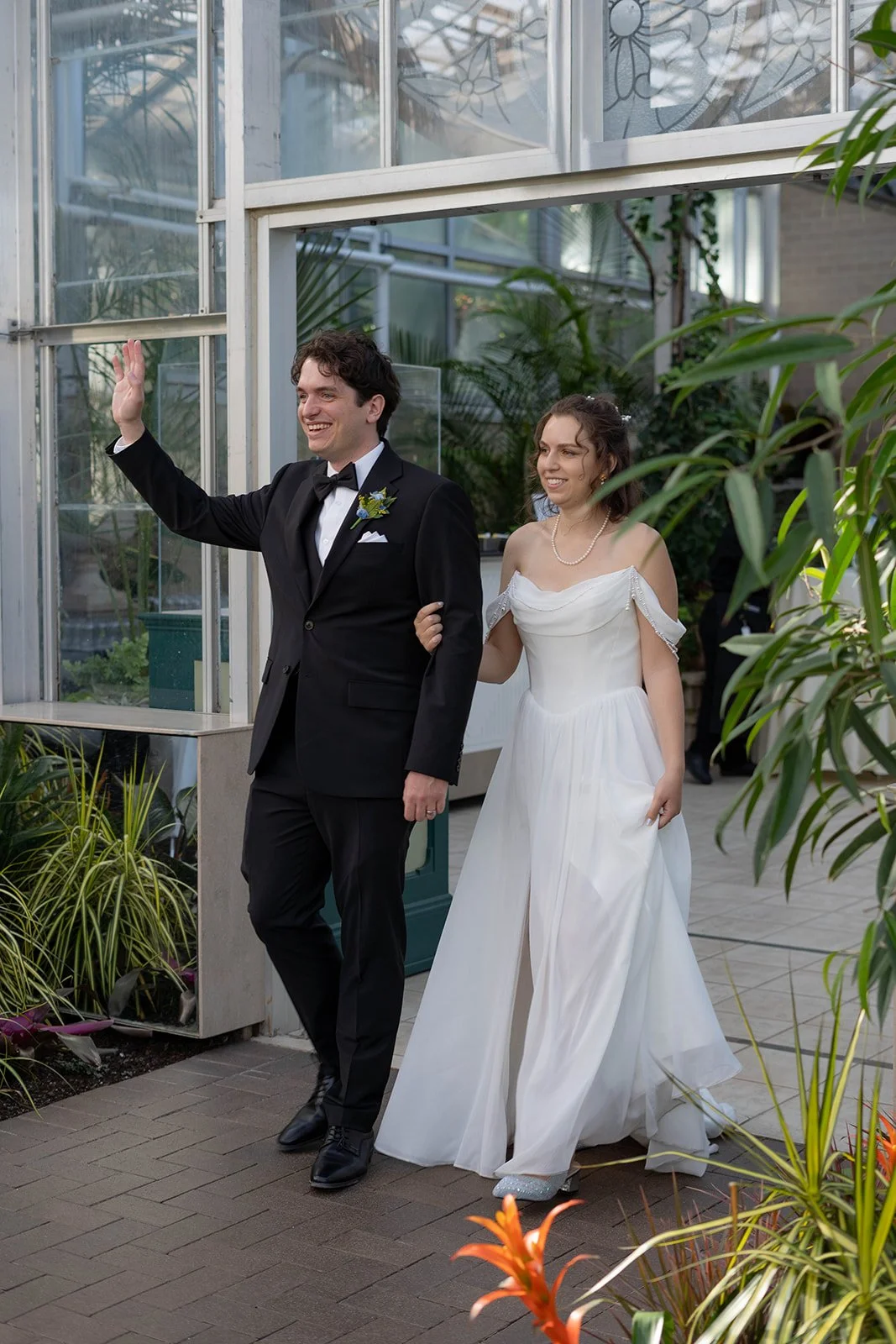 A newlywed couple, the groom in a black tuxedo and the bride in a white wedding dress, walks arm-in-arm through a greenhouse or conservatory, smiling and waving.