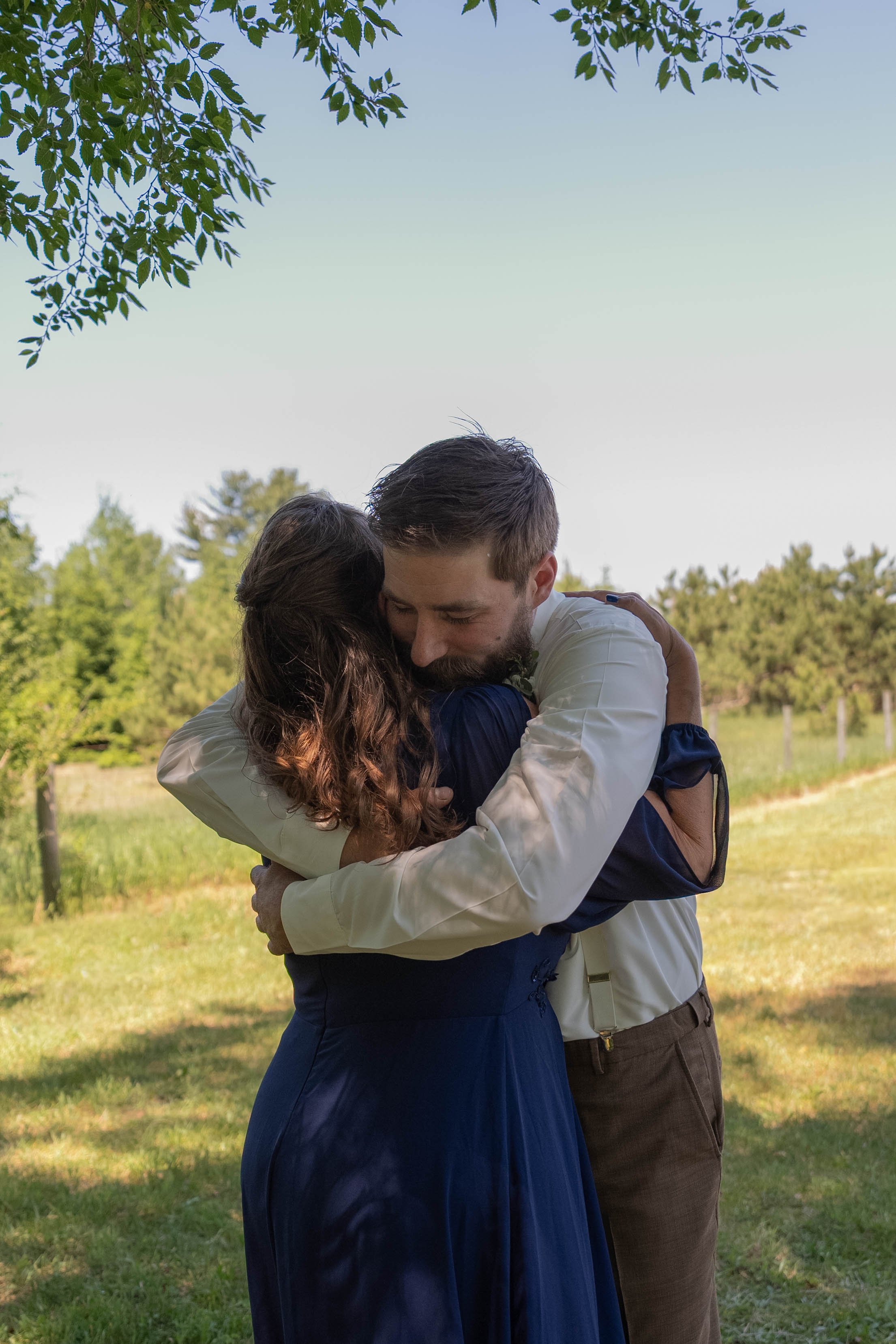 A man and woman embrace outdoors in a park or field, with trees and a clear sky in the background.