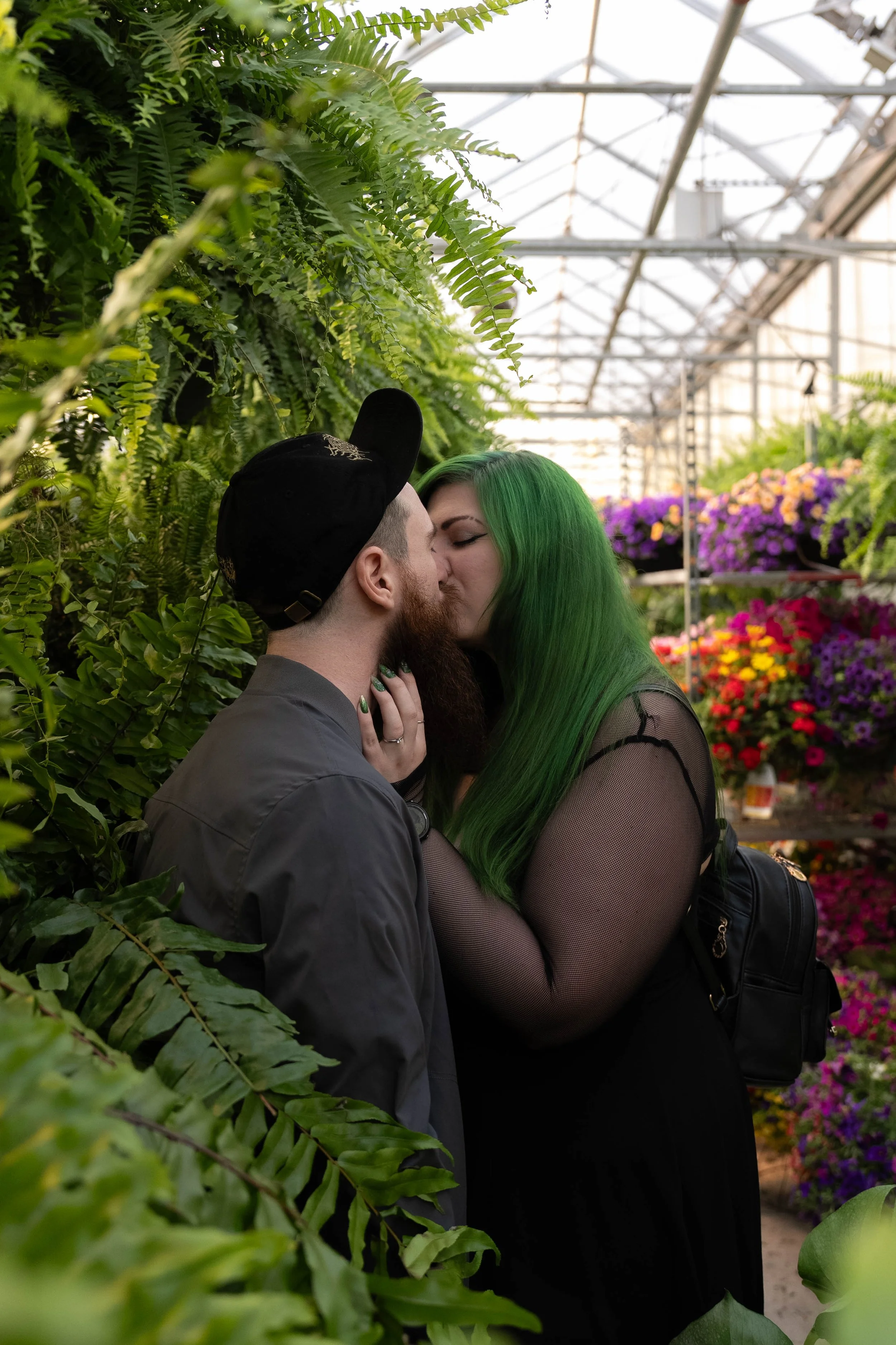A couple passionately kiss in a greenhouse filled with lush green plants and colorful flowers.