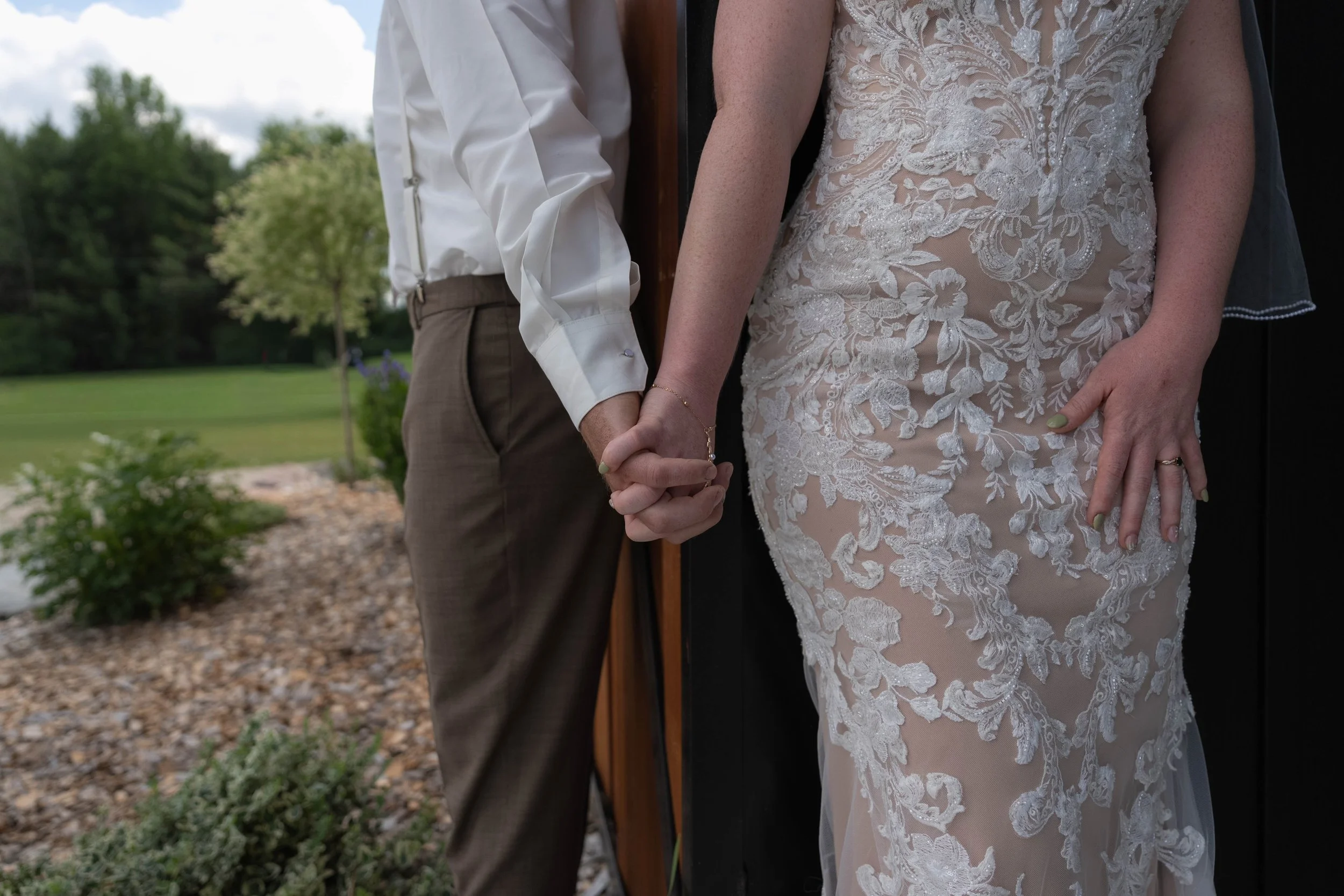 A bride and groom holding hands, with the bride in a lace wedding dress and the groom in a white shirt and brown trousers, standing outdoors near a wooden post.