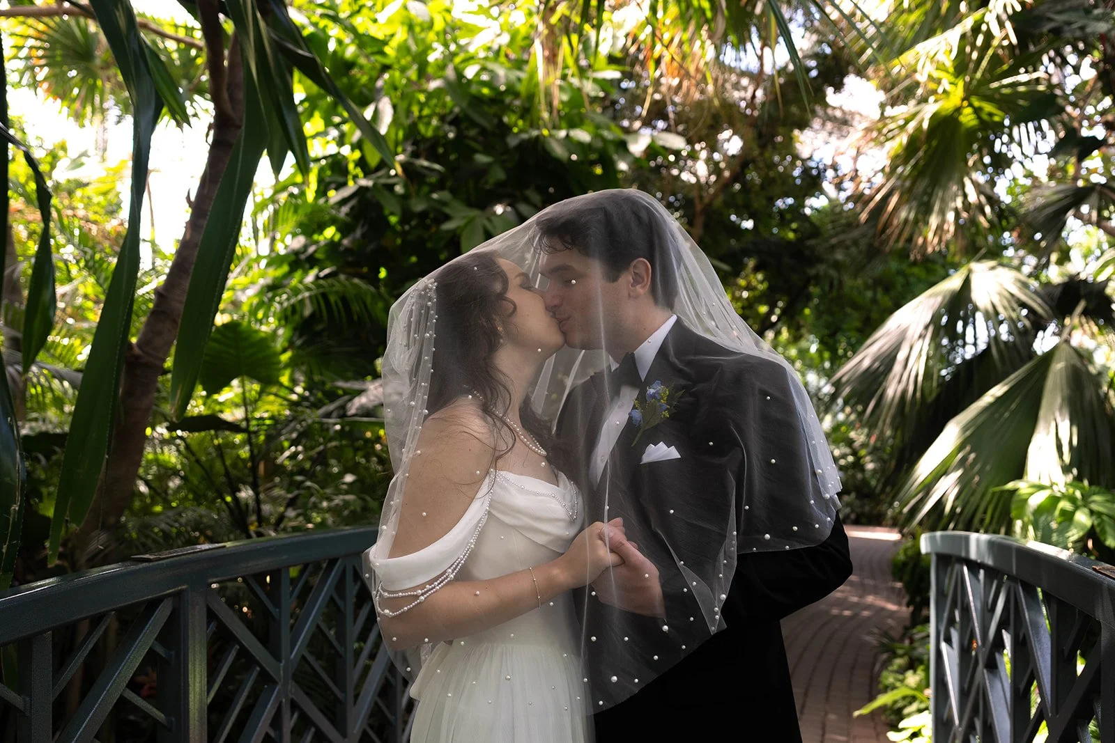 A bride and groom sharing a kiss under a veil in a lush, green outdoor setting with tropical plants.