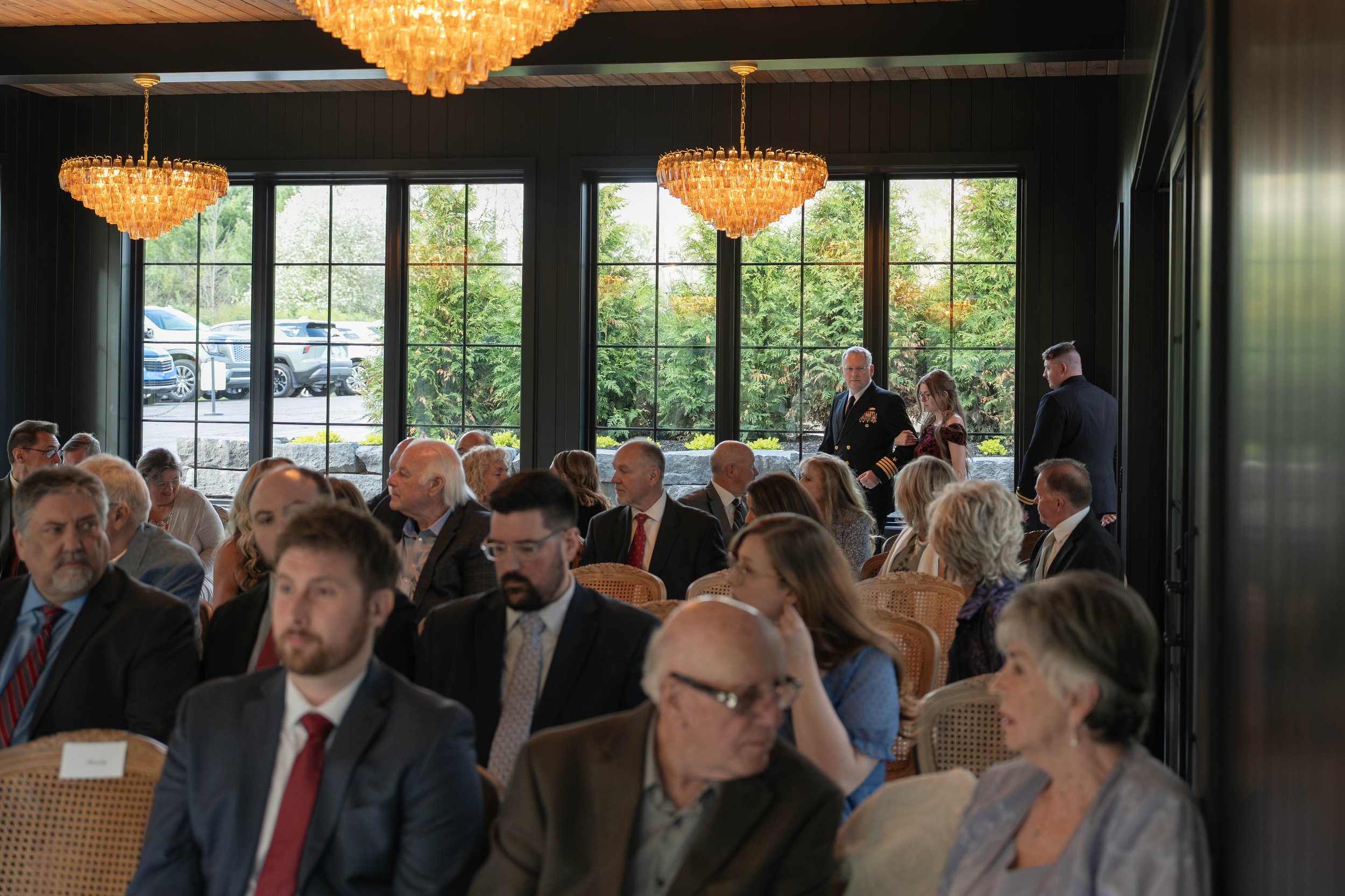 People attending a formal event or conference in a room with large windows, dark walls, and elegant chandeliers.