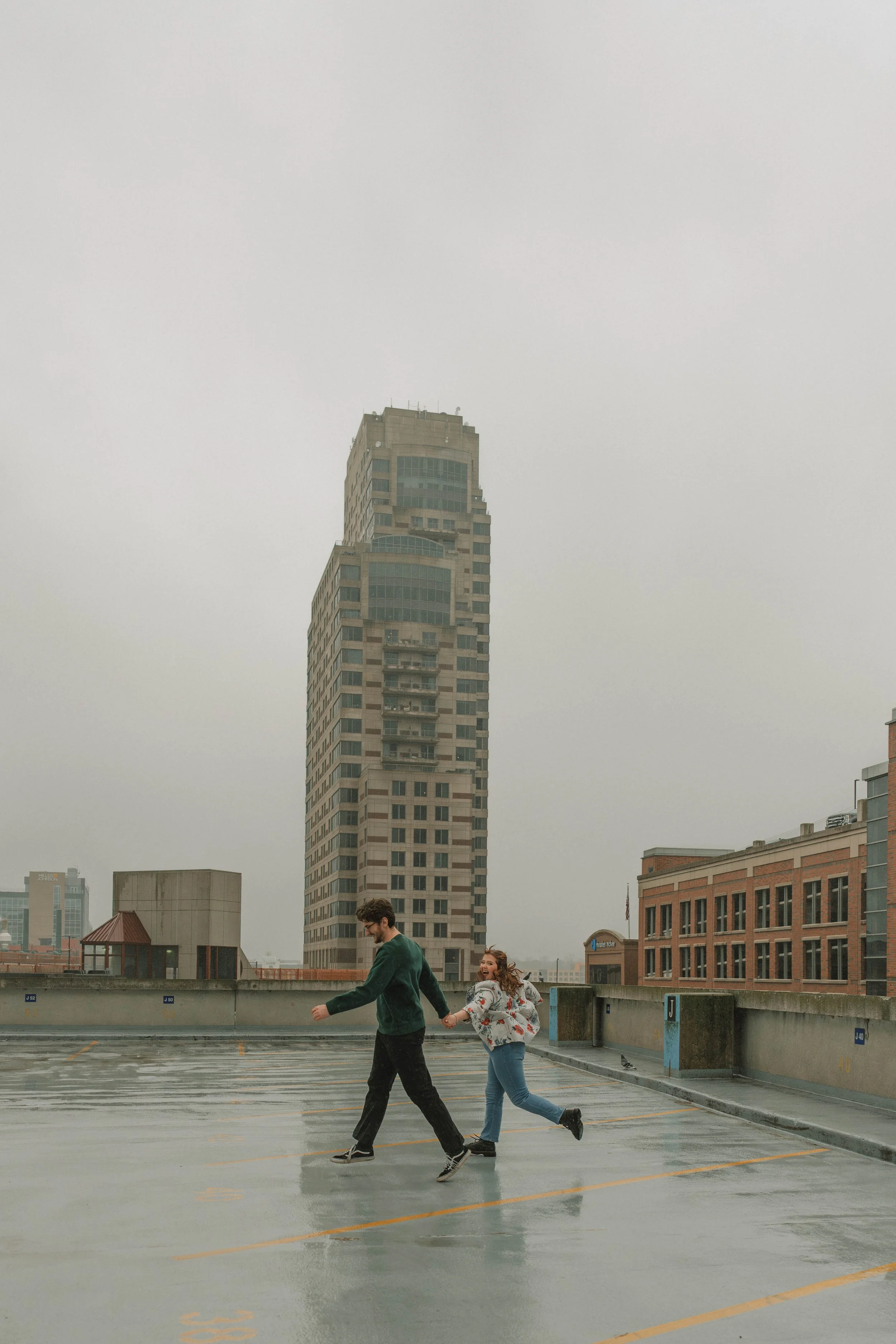 A man and a woman holding hands while walking in a parking garage on a rainy day. The city skyline with tall buildings is visible in the background.
