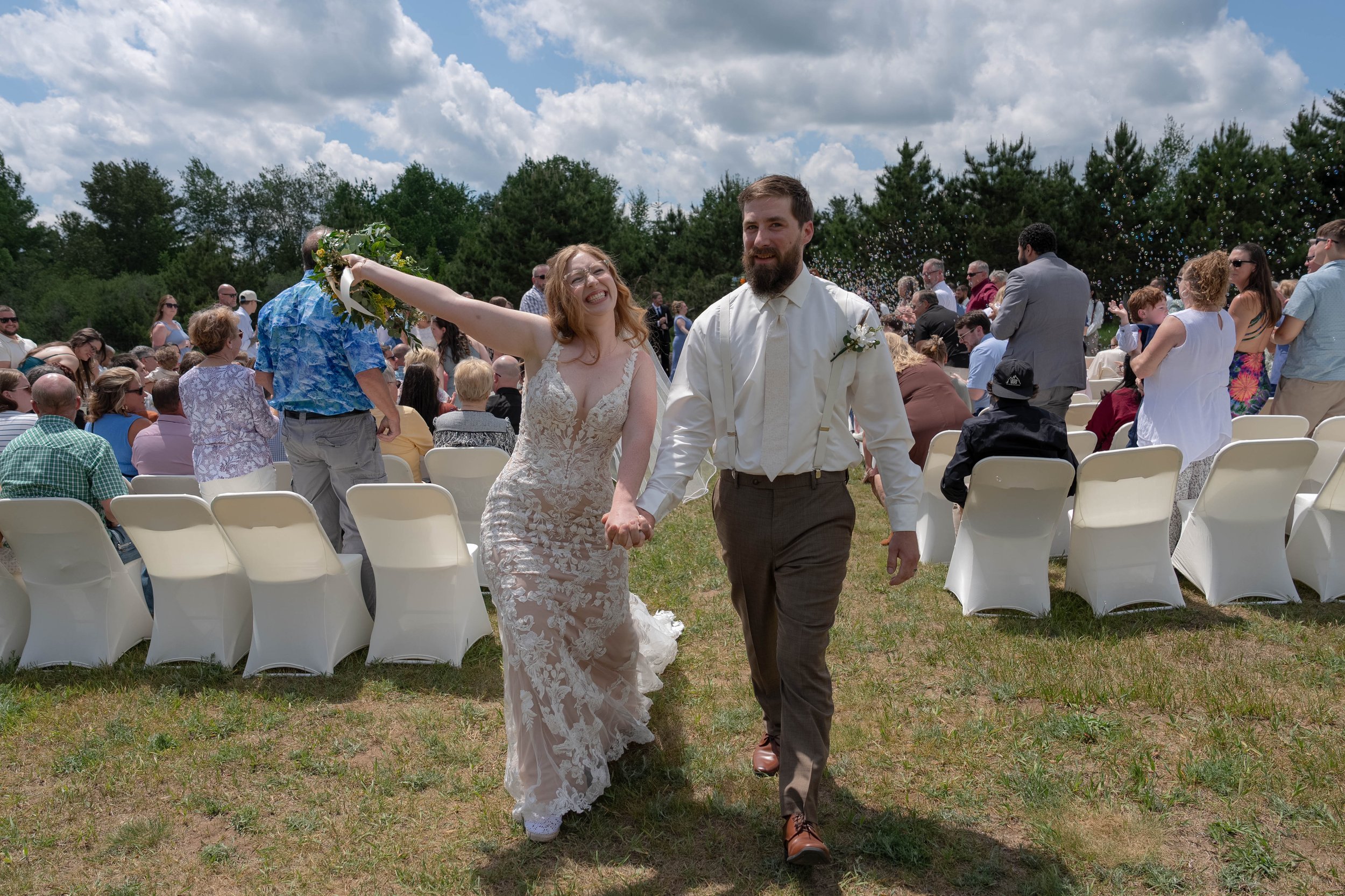 A bride and groom holding hands and smiling as they walk through a wedding ceremony outdoors, surrounded by seated guests on white chairs and a line of trees in the background under a partly cloudy sky.