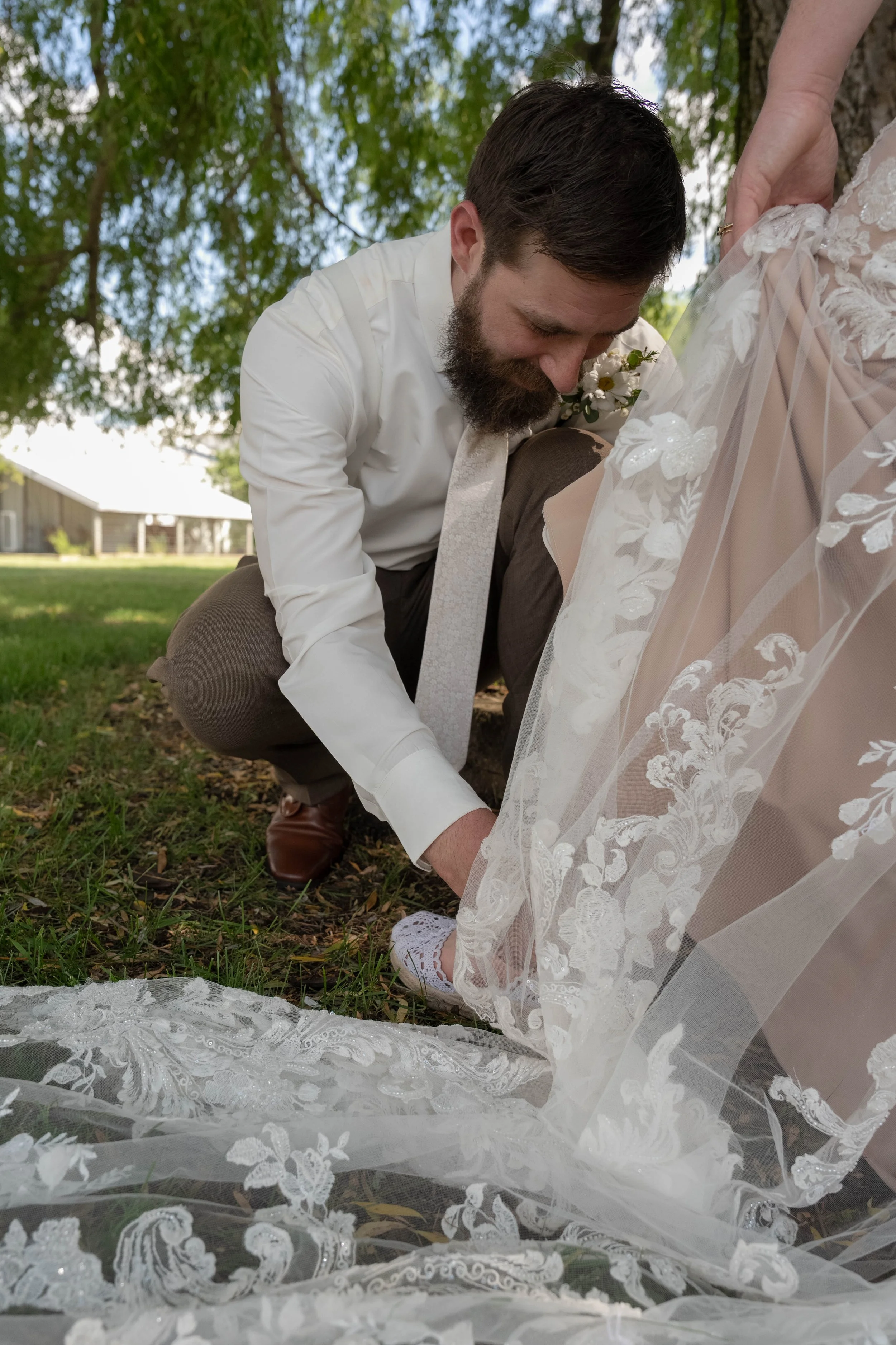 A man with a beard dressed in a white shirt and tie kneeling on the ground, tying the train of a bride's lace wedding dress outdoors under a tree.