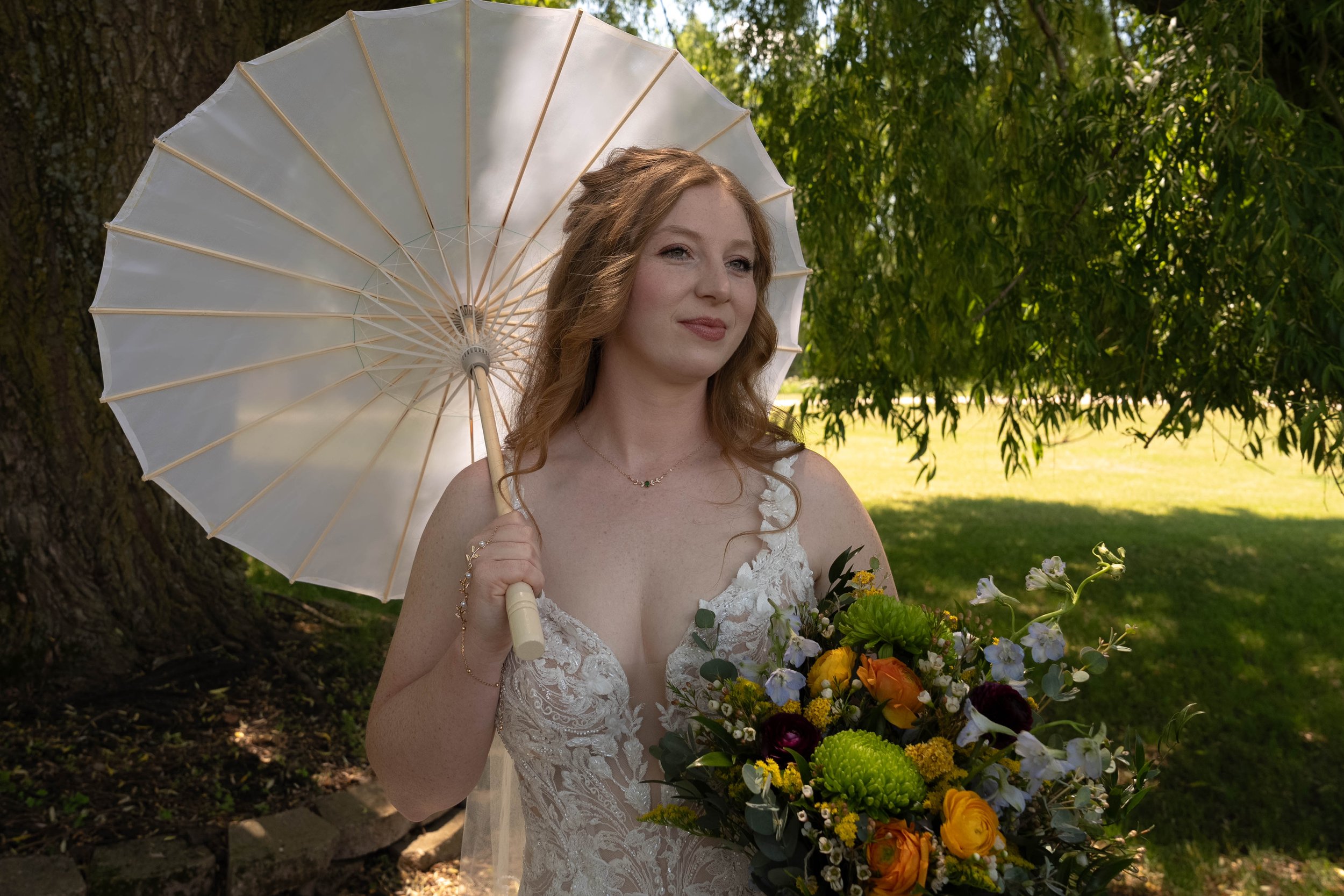 A bride standing outdoors holding a white parasol and a bouquet of colorful flowers, dressed in a white lace wedding gown, with greenery and trees in the background.