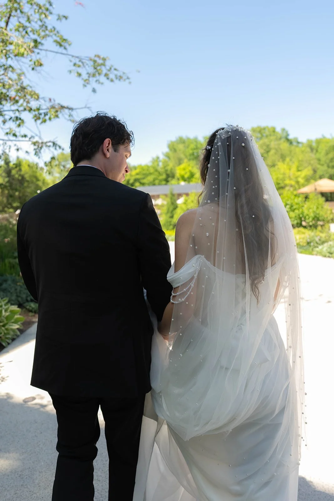 A bride and groom standing outdoors during a wedding, facing away from the camera, surrounded by greenery on a sunny day.