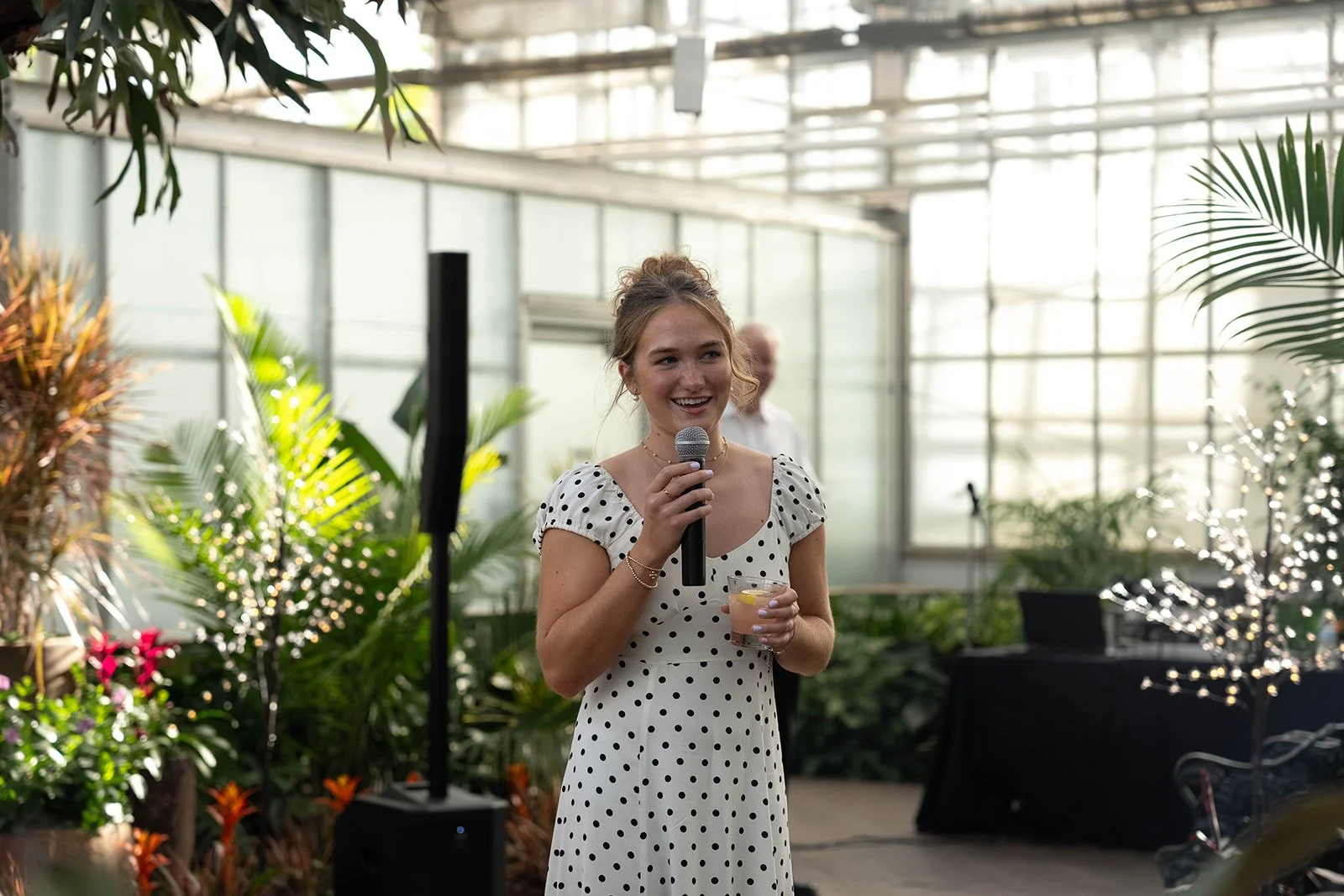 A young woman in a white dress with black polka dots holding a microphone and a clear cup with a lemon slice, smiling at an indoor event in a greenhouse or botanical setting.