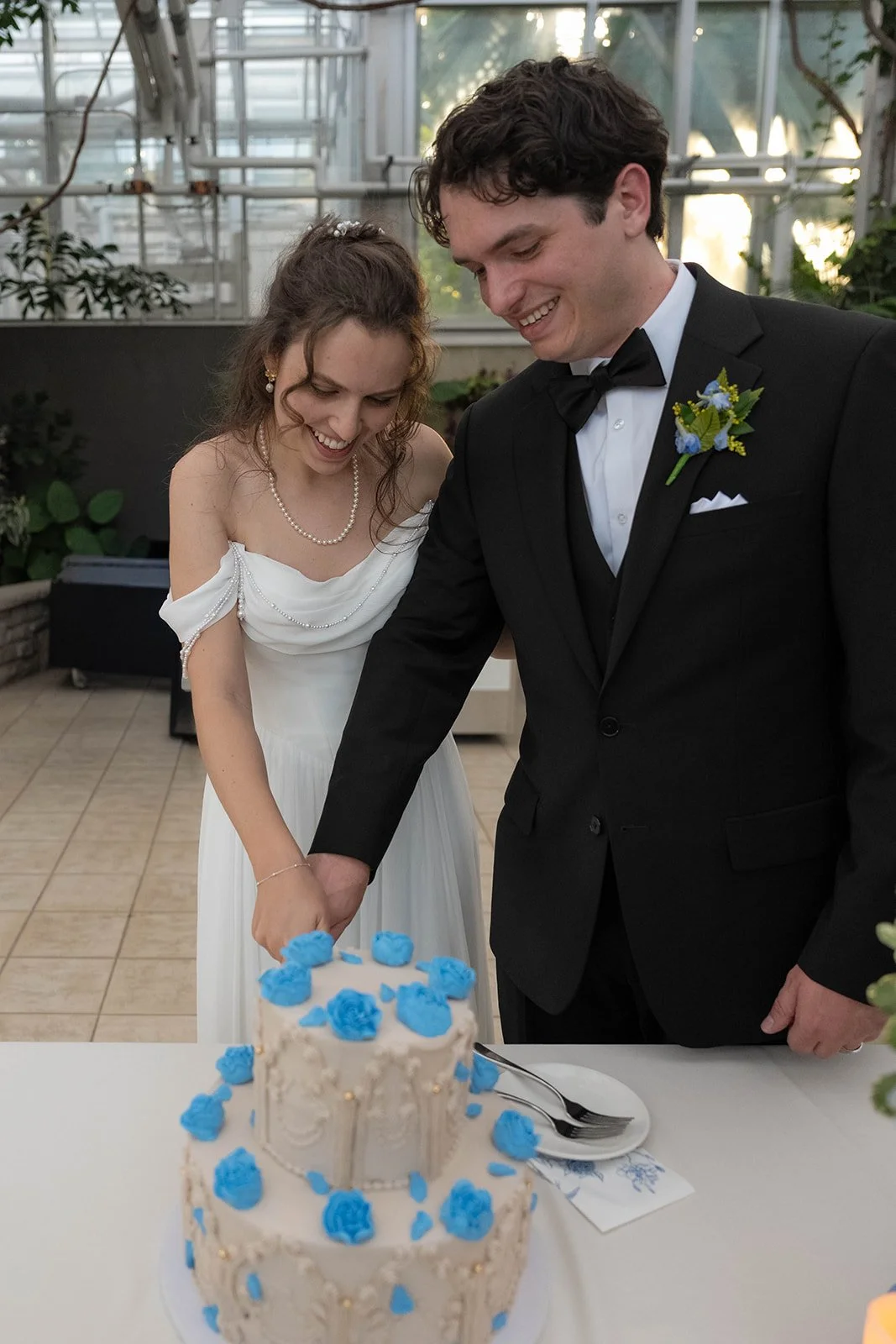 A bride and groom cutting a wedding cake together indoors. The bride is wearing a white dress with pearl details and a pearl necklace, while the groom is in a black tuxedo with a boutonniere. The wedding cake is decorated with blue roses.