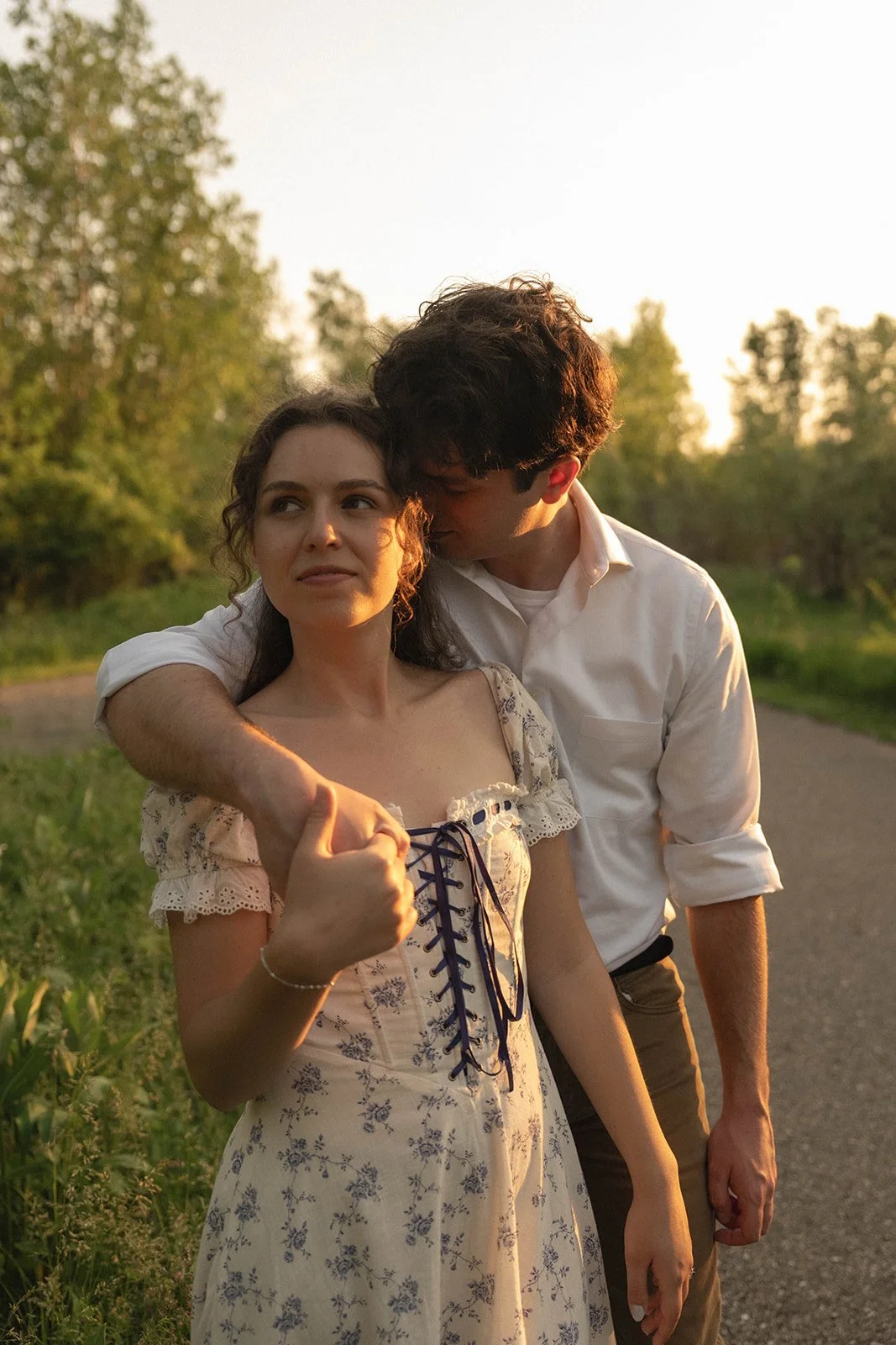 A young man and woman walking on a rural path during sunset, with the man embracing the woman from behind and kissing her on the head. The woman is looking to the side with a slight smile, wearing a floral dress, while the man is wearing a white shir