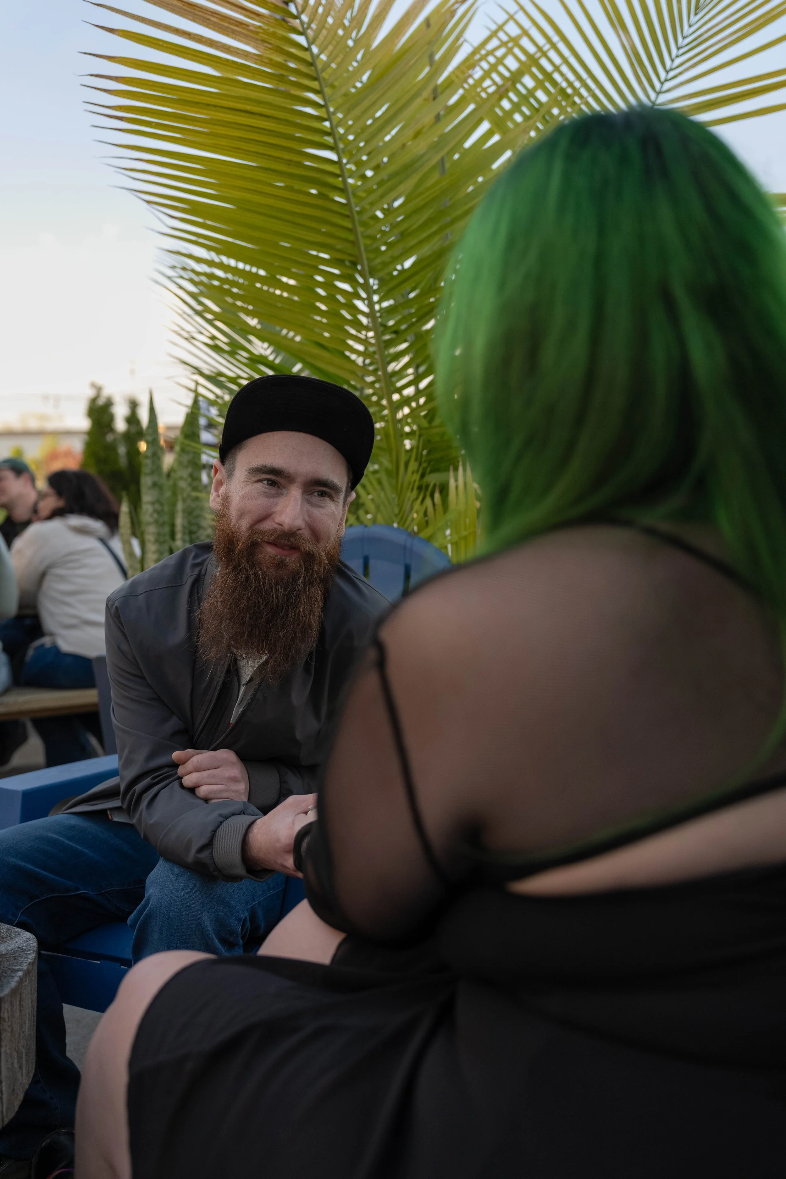 A man with a beard and black cap smiling and sitting outdoors, with large green palm leaves and a woman with green hair in the foreground, and other people in the background.