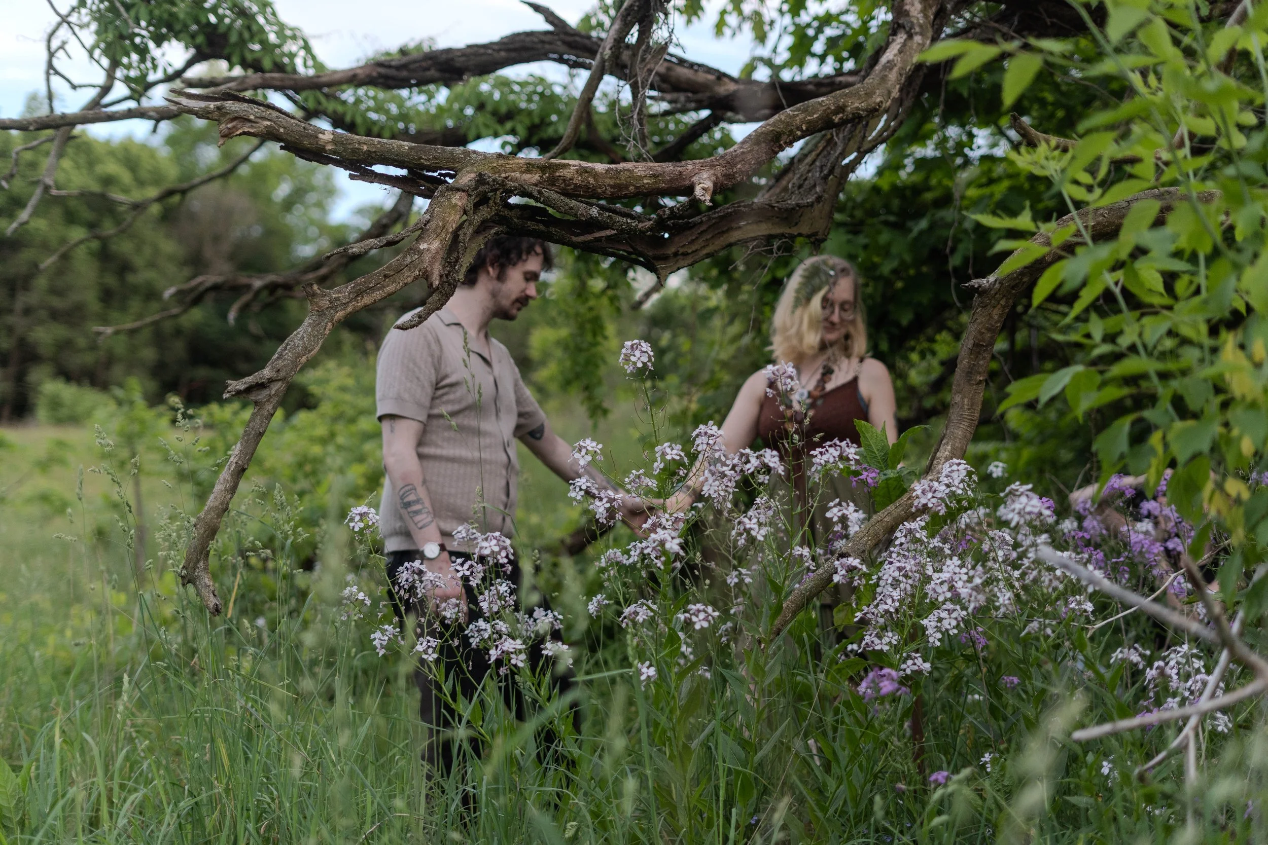 Two people, a man and a woman, standing in a lush green garden with purple flowers and surrounded by trees. The photo is taken from a low angle through grass and branches.